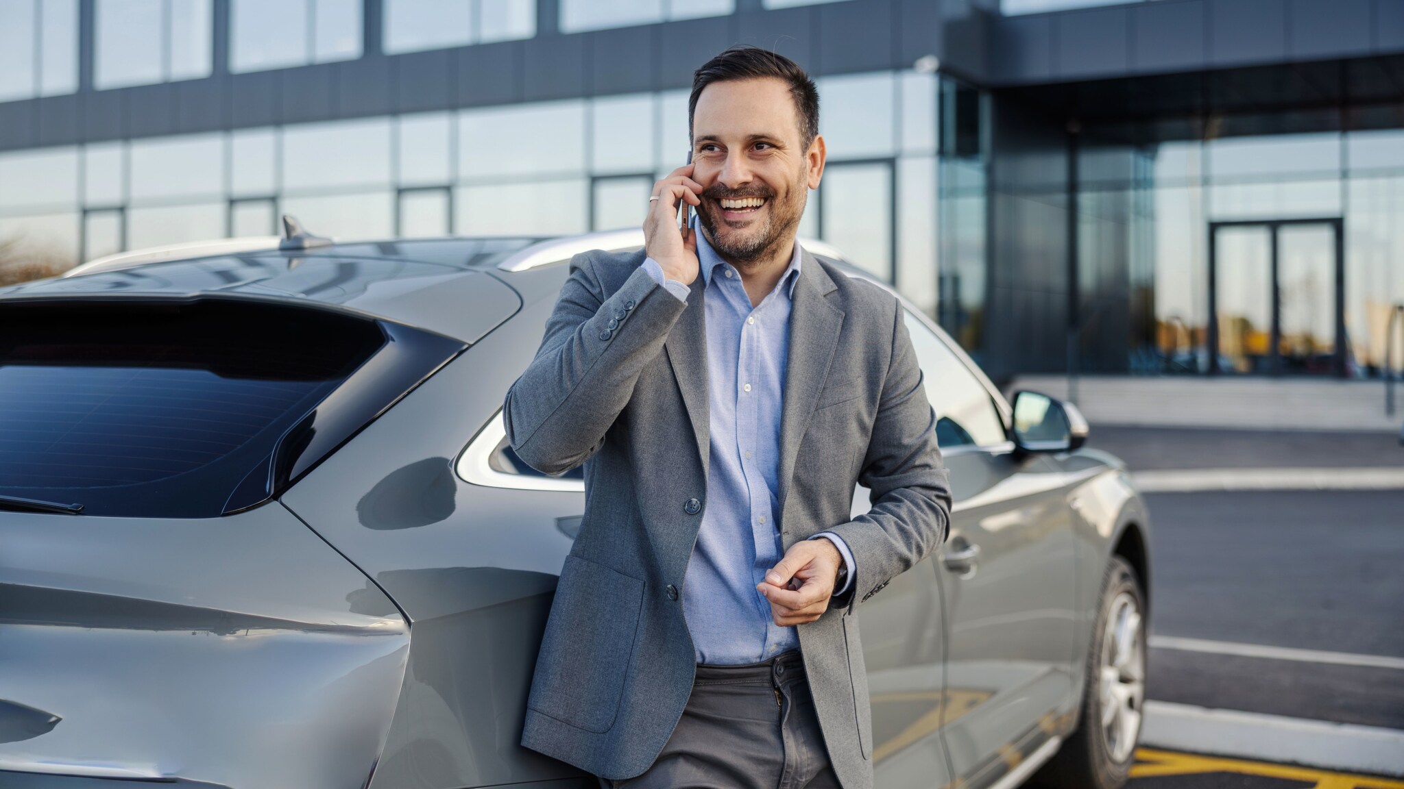Ein gut gelaunter Geschäftsmann im grauen Anzug lehnt an einem grauen Auto, während er ein Smartphone an sein Ohr hält auf einem Parkplatz vor einem Bürogebäude. 