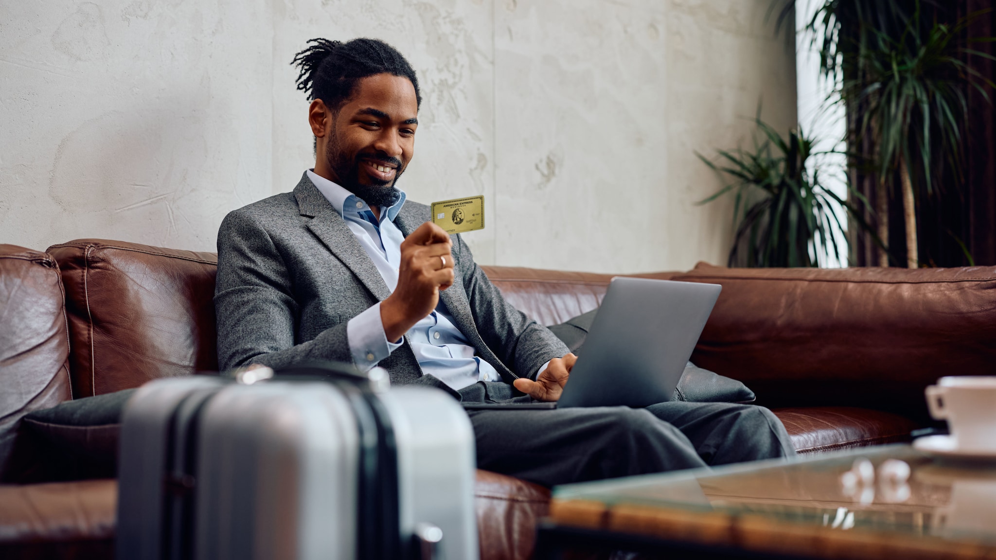amex-sbs-american-express-gold-business-versicherung-mann-laptop Ein Geschäftsmann im grauen Anzug sitzt mit Laptop auf dem Schoß auf einem Sofa in einer Lounge und hält er eine goldene Business Kreditkarte von American Express in der Hand hoch, im Vordergrund steht ein Koffer.