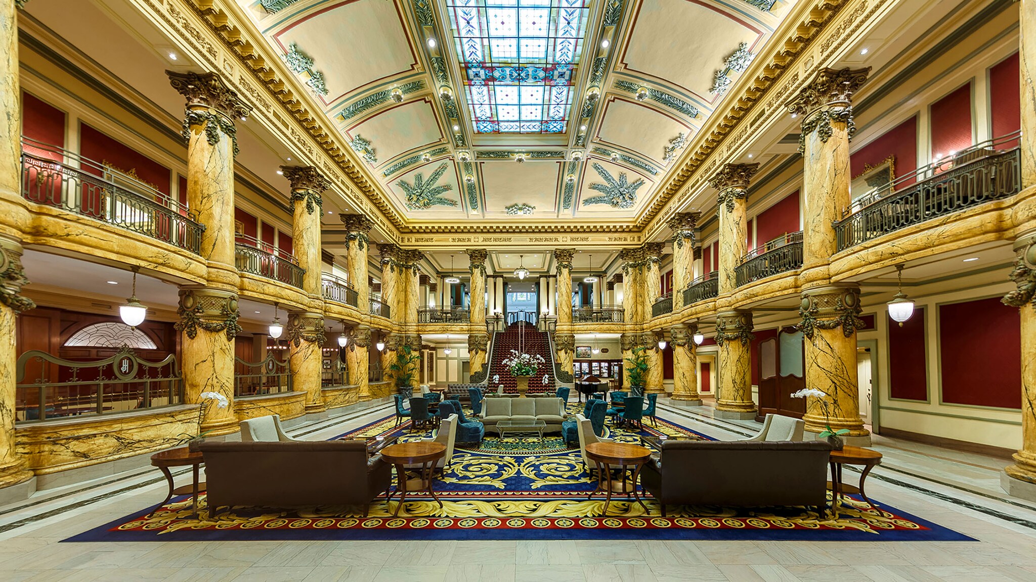In einer großen Hotel-Lobby stehen, umgeben von verzierten Säulen, Sessel und Tische auf einem großen, bunten Teppich. 