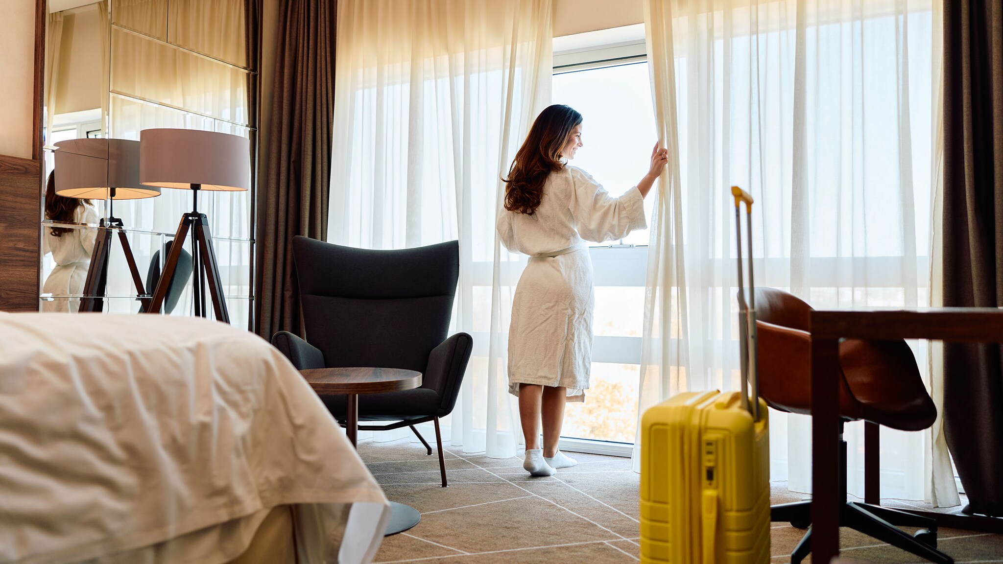 Rückansicht einer Frau im weißen Bademantel, die an einem Fenster mit weißen Gardinen in einem Hotelzimmer mit Koffer steht.