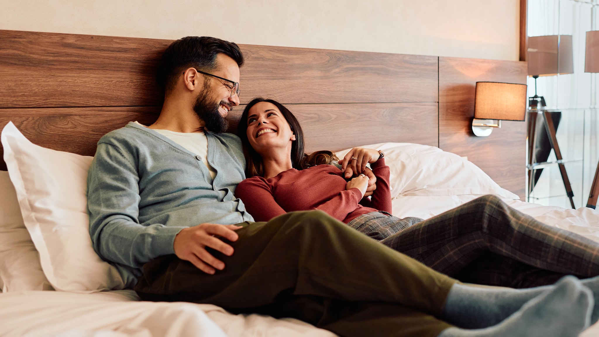 Amex Hotels Ein Mann und eine Frau liegen in Alltagskleidung Arm in Arm in einem Hotelbett und schauen sich lächelnd an.