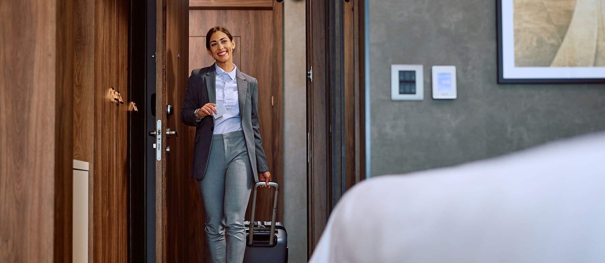 Amex Hotels Eine lächelnde Frau im Business-Outfit mit Trolley und Schlüsselkarte in den Händen betritt ein Hotelzimmer, im Vordergrund ein Bett im Anschnitt.