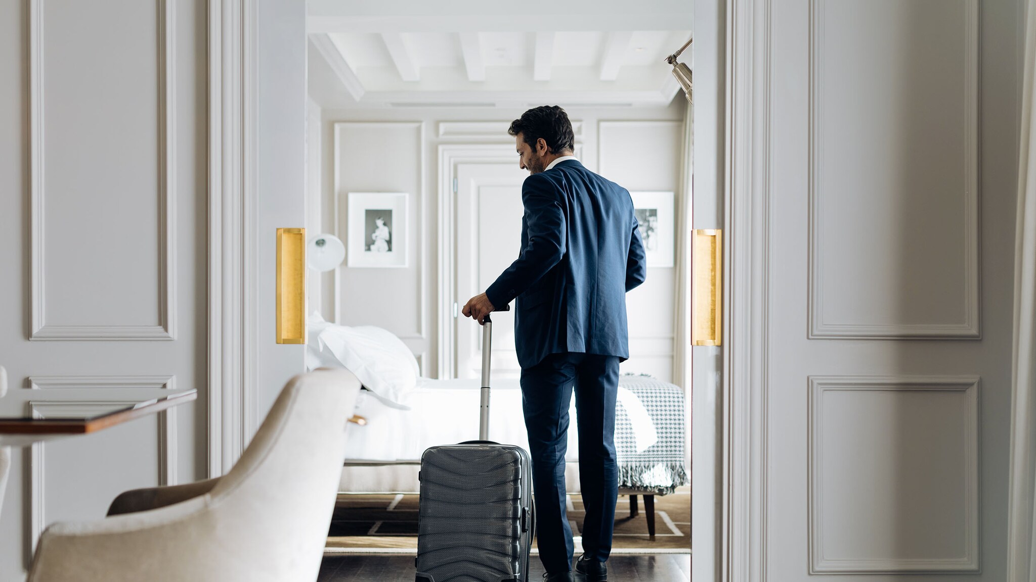 Rückansicht eines Mannes im blauen Anzug, der mit einem Trolley das Schlafzimmer einer hellen Hotelsuite betritt.