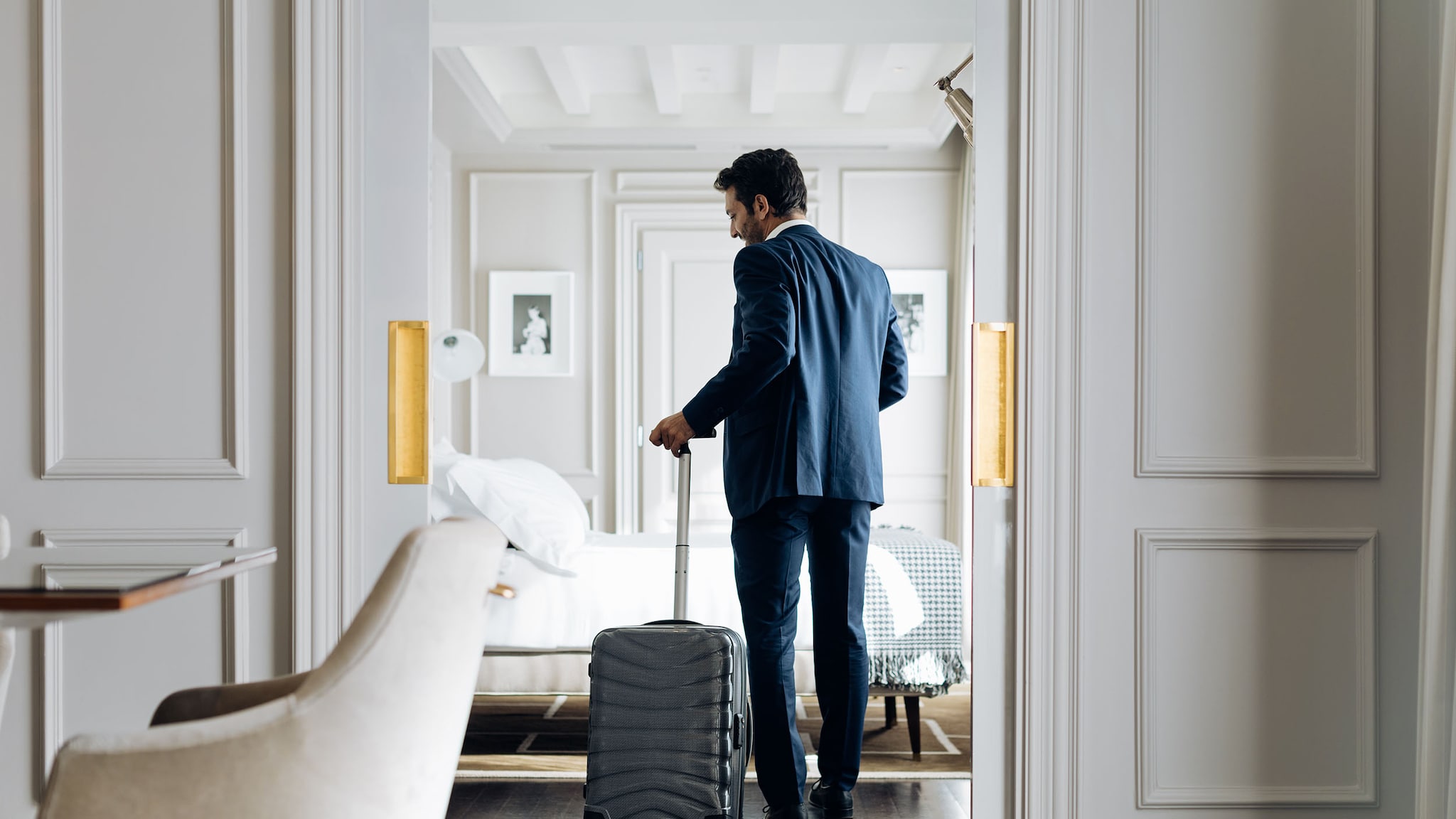 Amex Hotels Rückansicht eines Mannes im blauen Anzug, der mit einem Trolley das Schlafzimmer einer hellen Hotelsuite betritt.