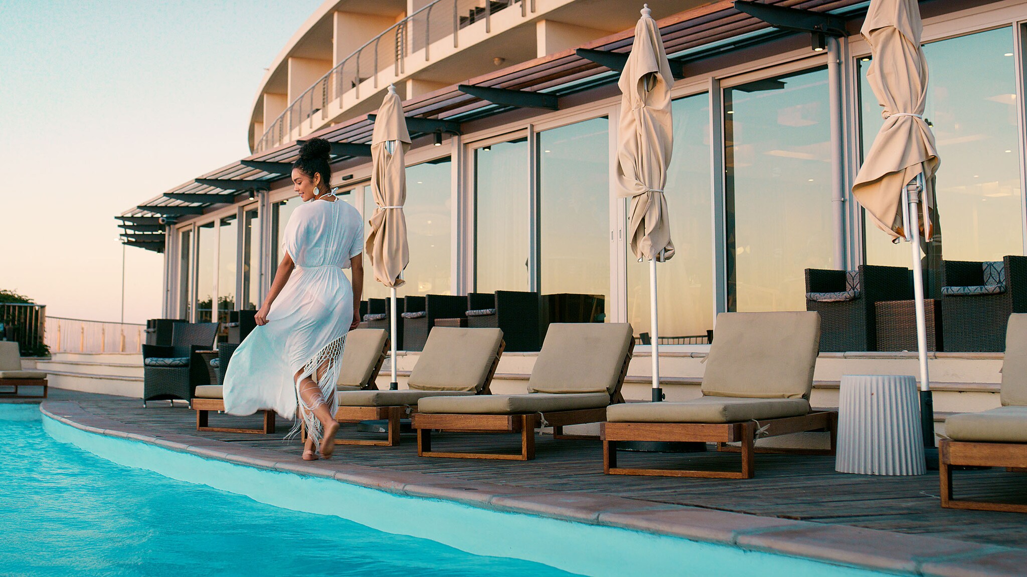 Rückansicht einer Frau im weißen Sommerkleid, die auf einer Hotelterrasse an einem Pool entlangläuft. 
