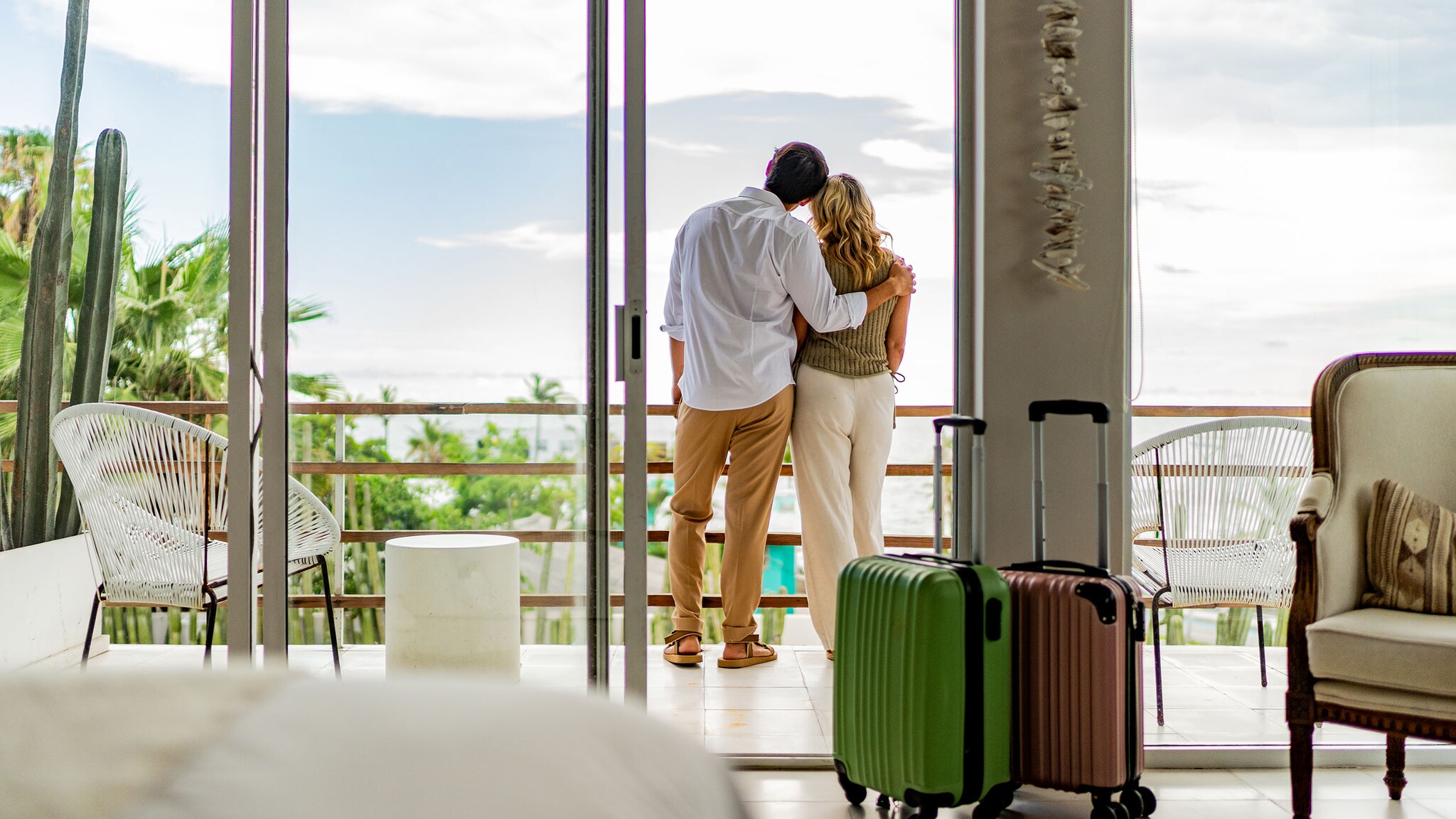 Rückansicht eines sich umarmenden Paares in Sommerkleidung auf dem Balkon eines Hotelzimmer vor tropischer Landschaft. 