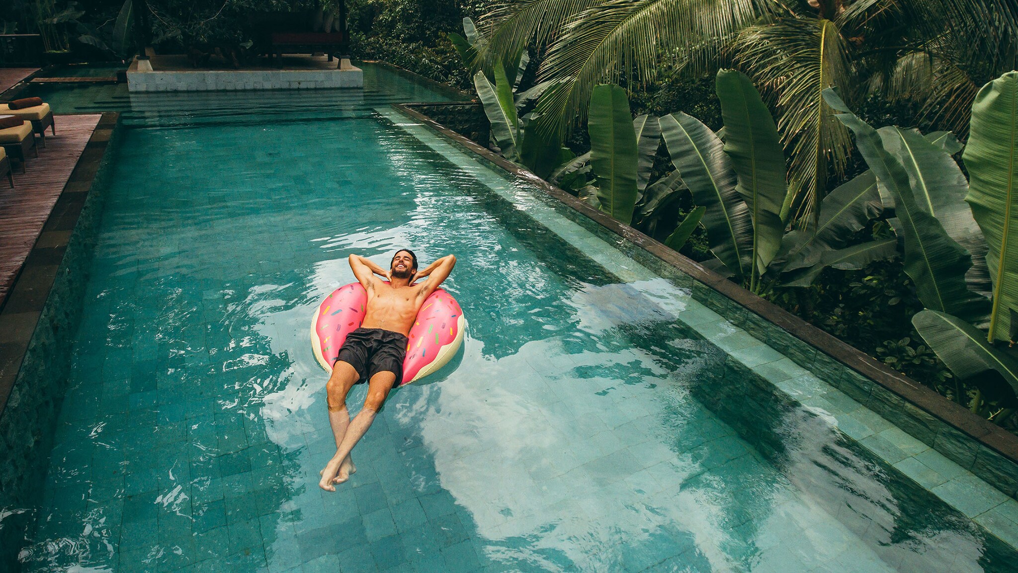 Ein Mann in Badehose treibt entspannt auf einem pinken Schwimmreifen im Wasser eines Hotelpools inmitten tropischer Vegetation.