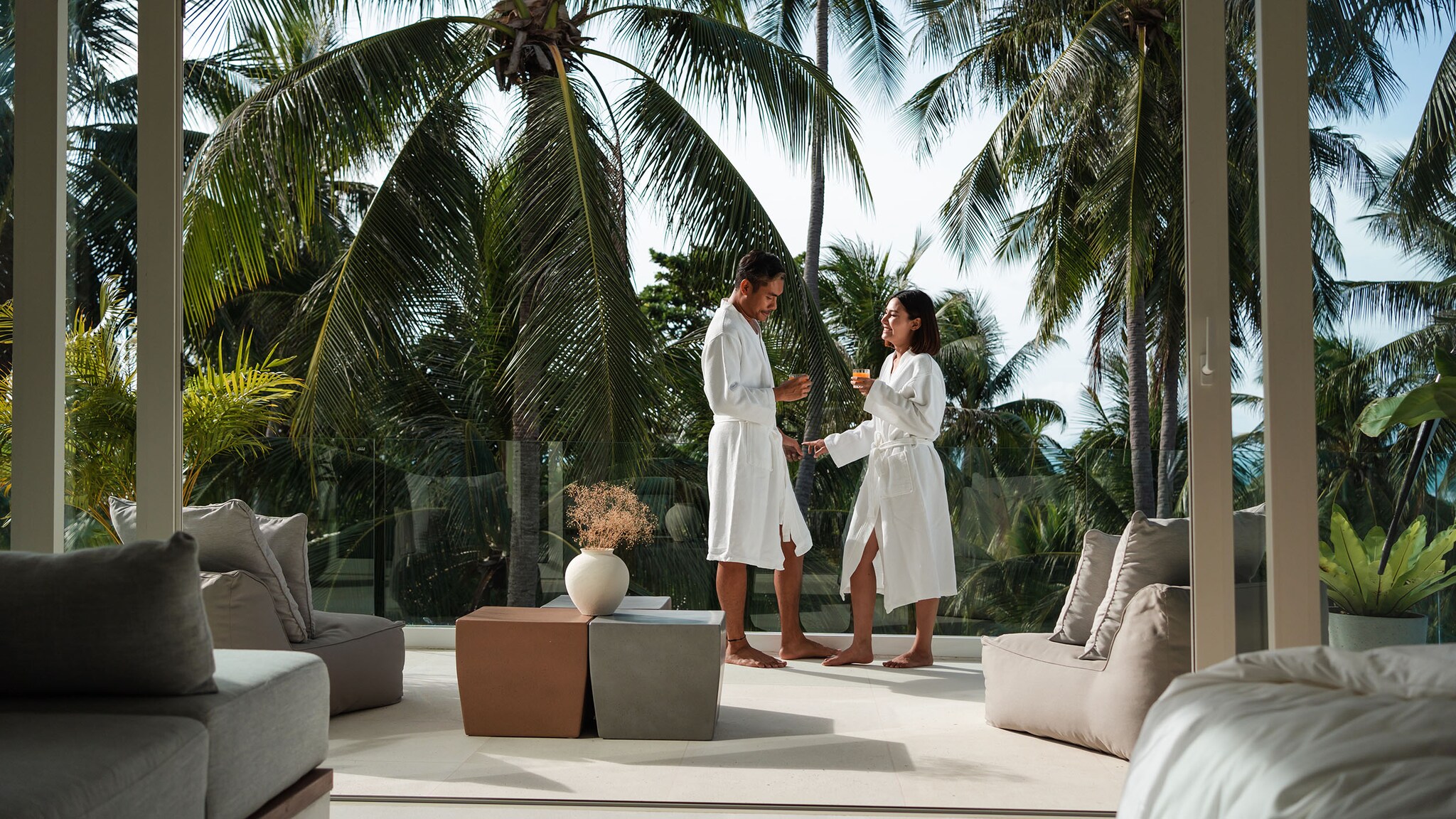 Ein Mann und eine Frau in weißen Bademänteln stehen sich gegenüber mit Getränken in den Händen auf einer Hotelterrasse mit Sitzgelegenheiten vor einem Palmenwald.