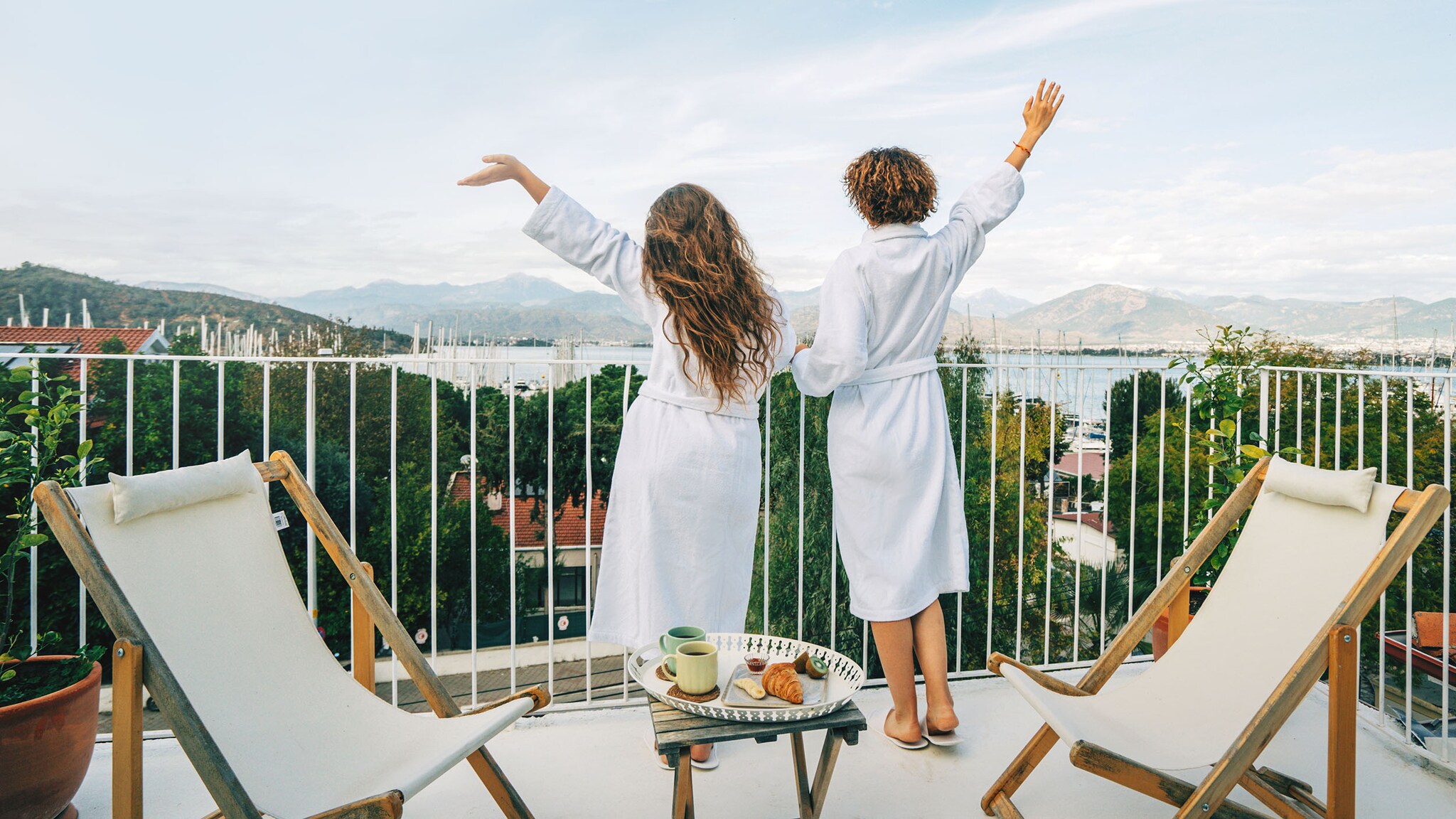 Rückansicht von zwei Personen in weißen Bademänteln, die mit ausgestreckten Armen von einem Balkon mit Seepanorama winken.