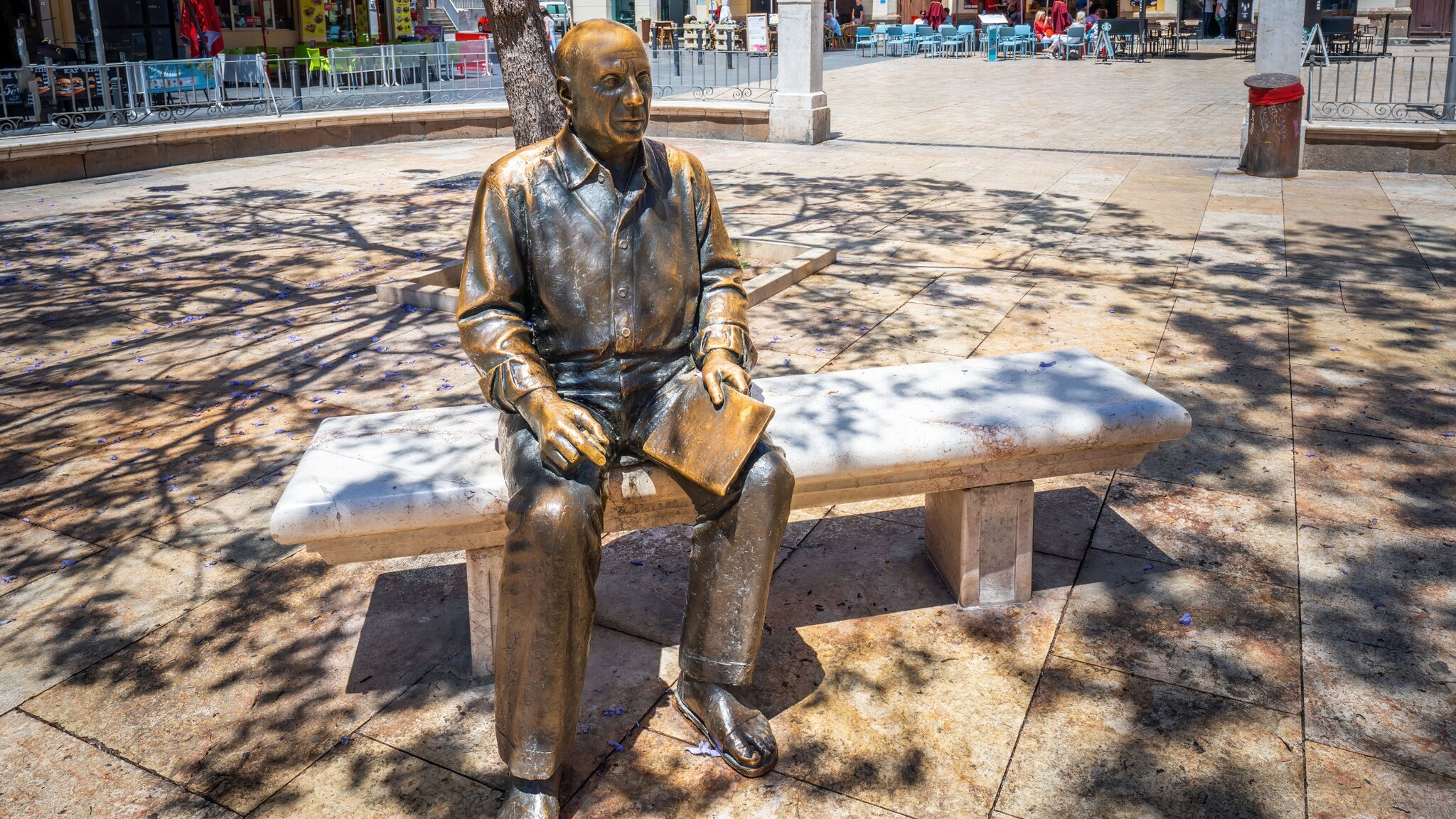 Bronzestatue von Picasso, auf einer Bank sitzend, auf einem Platz in einer Stadt.