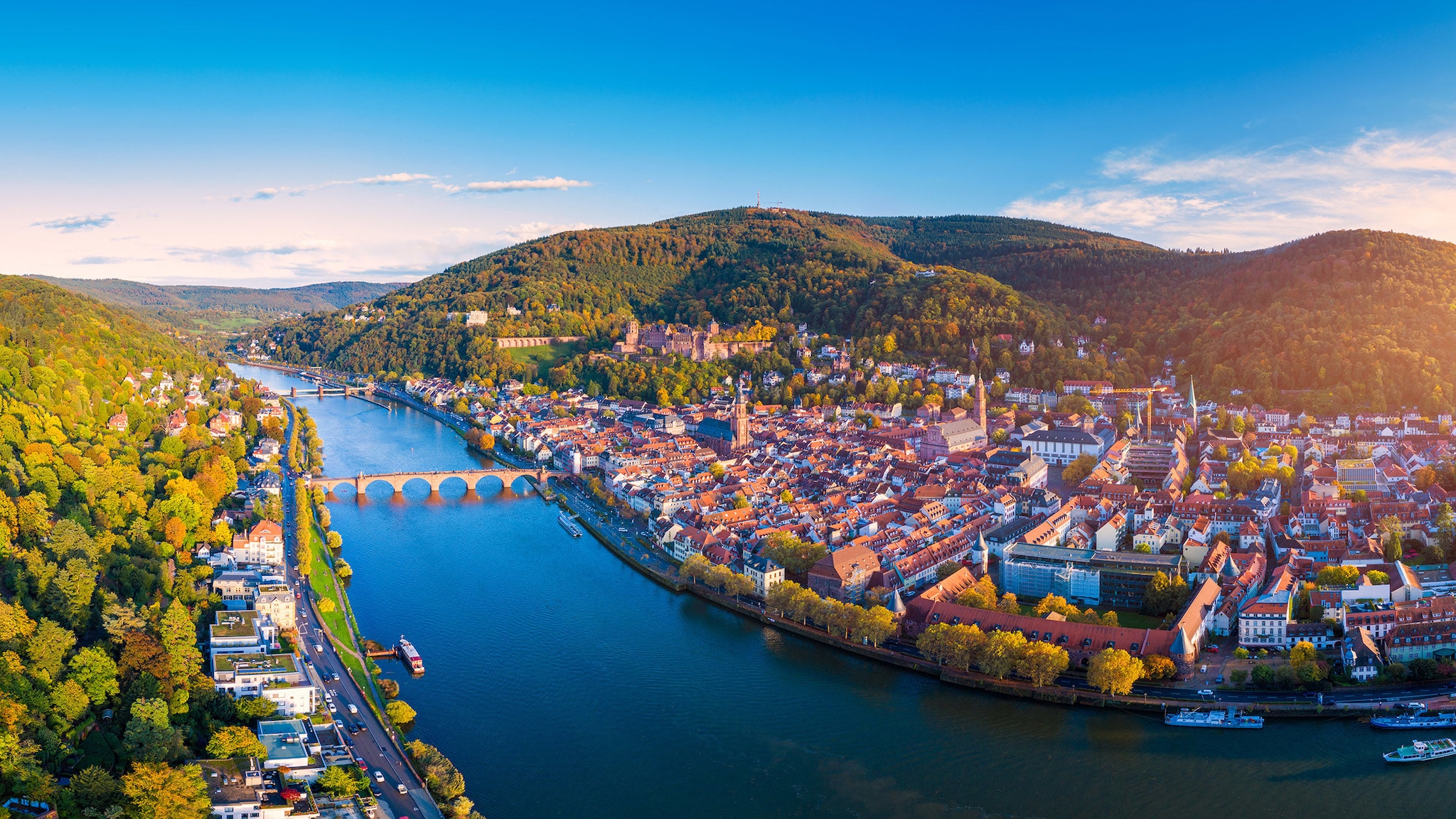 amex-magazin-schoensten-staedte-deutschlands-altstadt-fluss Luftaufnahme der Stadt Heidelberg mit historischer Altstadt am Fluss, umgeben von bewaldeter Hügellandschaft im warmen Sonnenlicht.