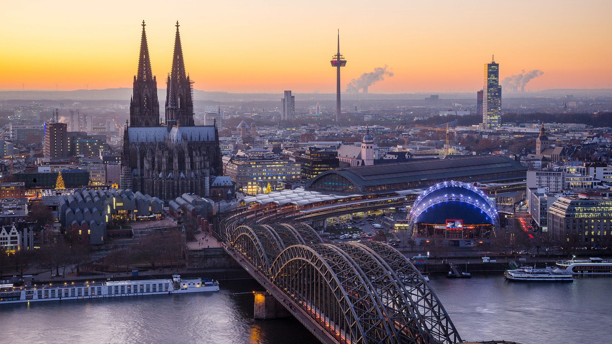 Luftaufnahme Stadtpanorama von Köln mit Dom und Eisenbahnbrücke am Fluss bei Sonnenuntergang.