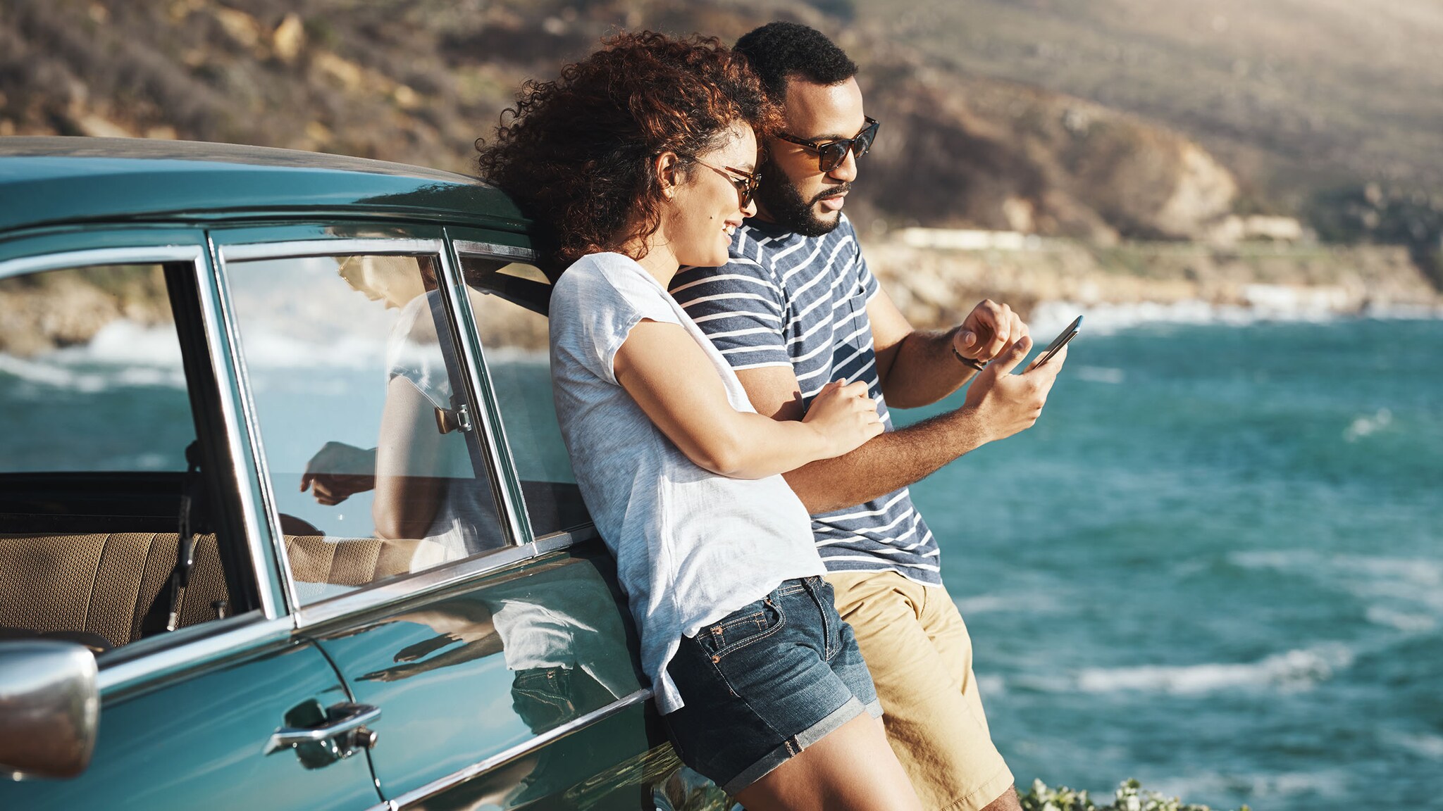 Ein Mann und eine Frau lehnen an einem Auto und schauen auf ein Smartphone.
