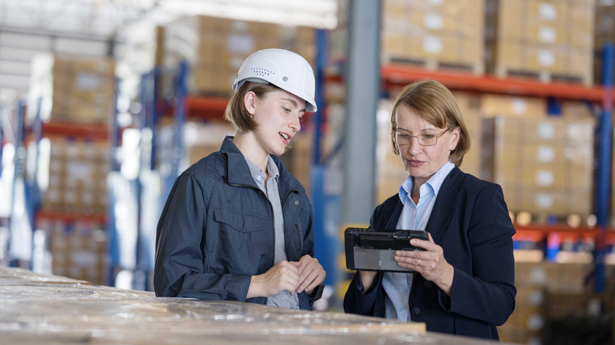 Corporate Card Vorteile Eine Frau mit Helm und eine Frau in Businesskleidung stehen in einem Lager und schauen sich etwas auf einem Tablet an
