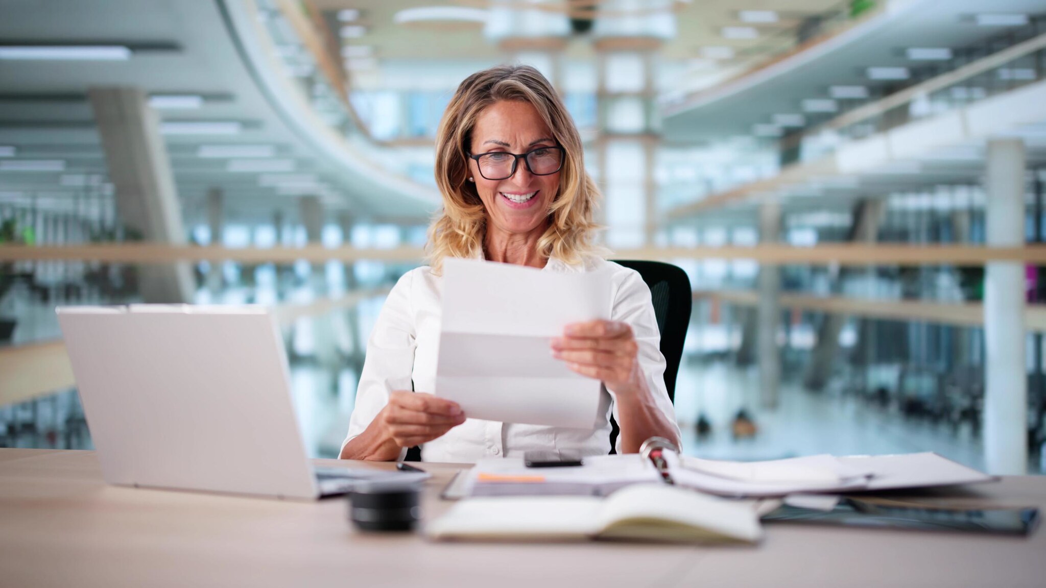 Eine Frau sitzt in einem lichtdurchfluteten Bürogebäude und schaut zufrieden lächelnd auf ein Dokument in ihren Händen