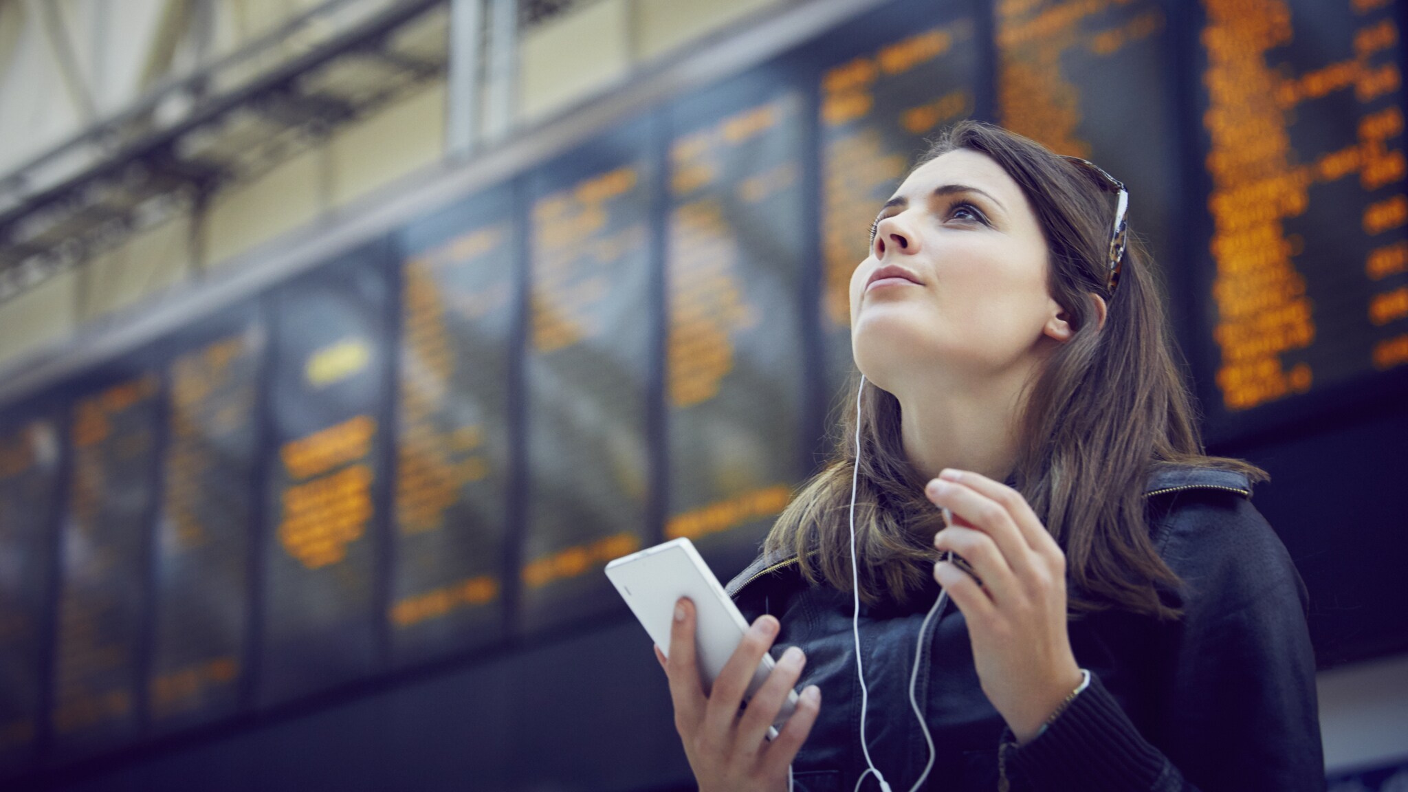 Eine junge Frau mit Smartphone in der Hand steht vor der digitalen Anzeigetafel eines Flughafens und schaut zuversichtlich nach oben