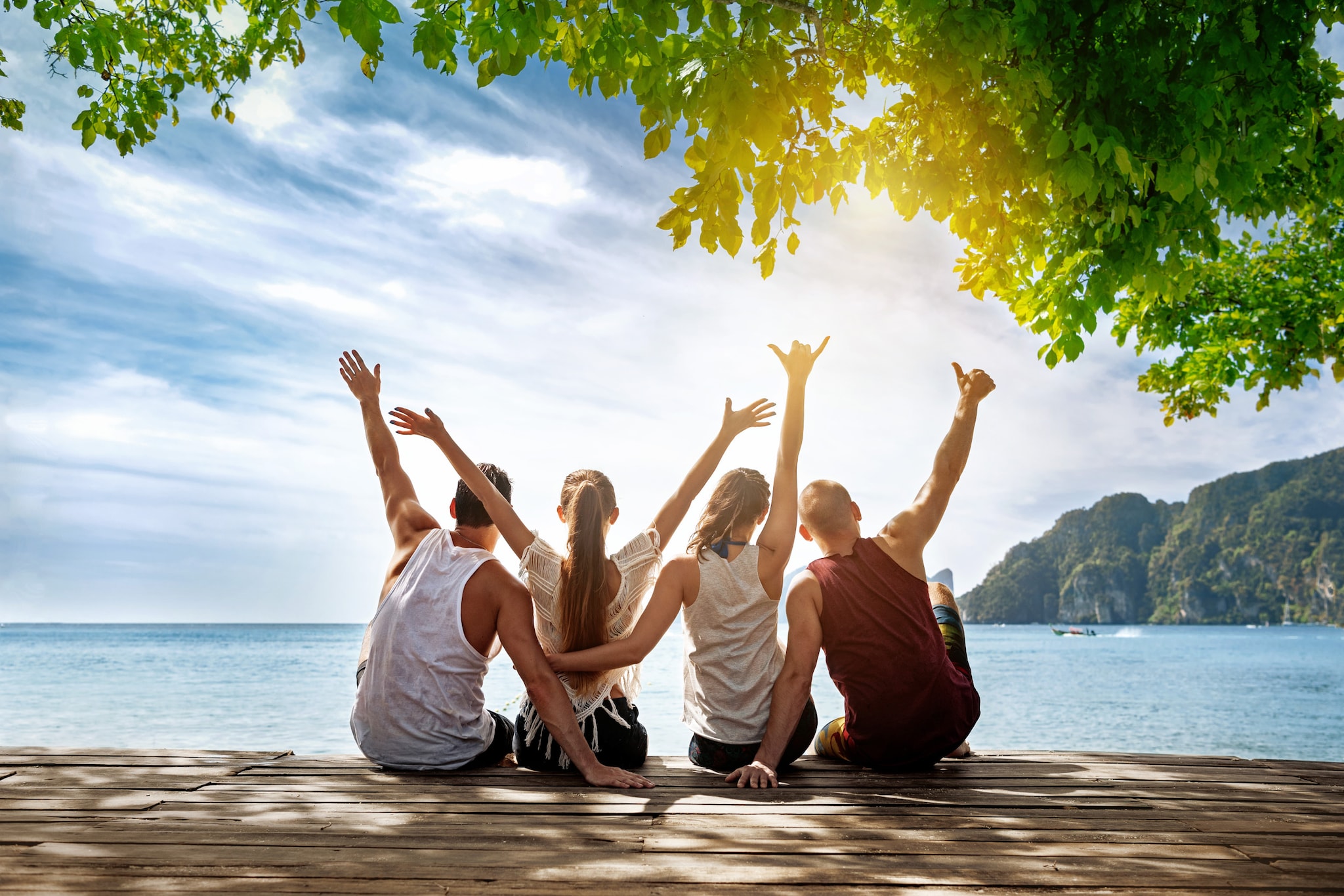 amex-insurances-reiseruecktrittsversicherung-gemeinsame-buchung-vier-personen-meer Vier Personen sitzen mit dem Rücken zur Kamera am Meer und halten ihre Hände in die Höhe.