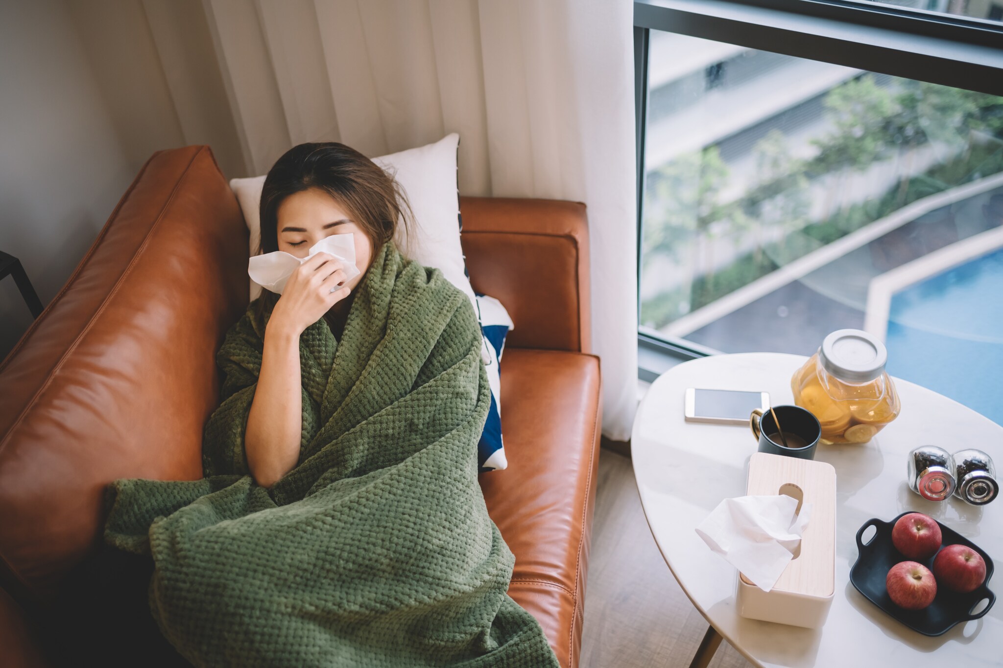 Eine Frau liegt, eingekuschelt in eine Decke, auf einem Sofa und putzt sich die Nase.