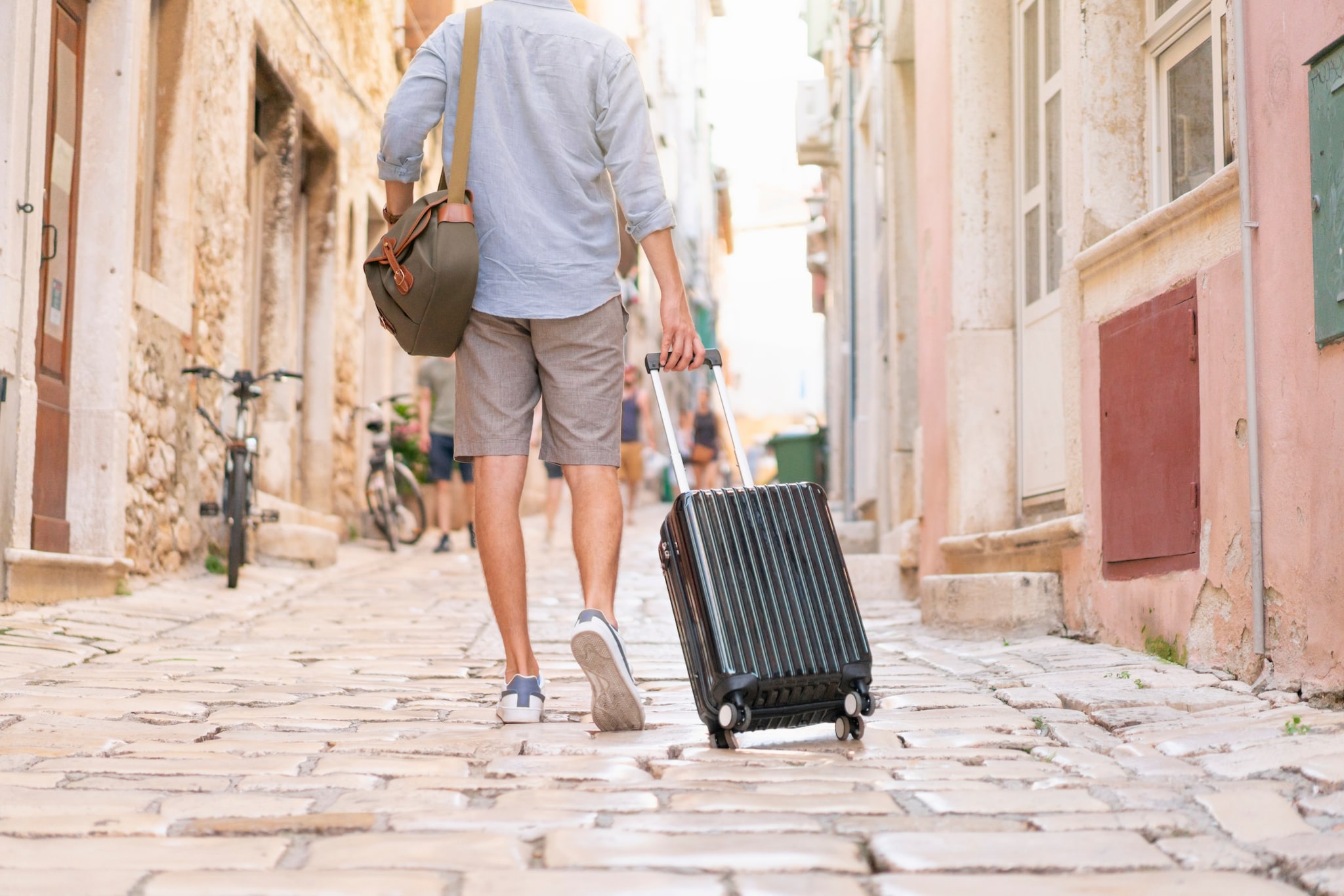 amex-insurances-diebstahl-ausland-versicherung-mann-koffer Mann in kurzen Hosen zieht seinen Rollkoffer über die Straße einer idyllischen mediterranen Altstadt.