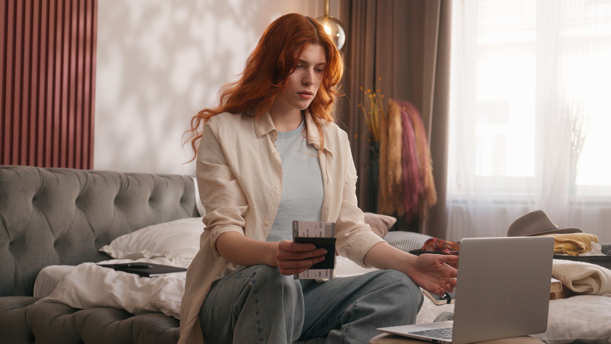 Besorgt blickende junge Frau sitzt auf einem Sofa vor ihrem Laptop und hält Flugtickets in der Hand.