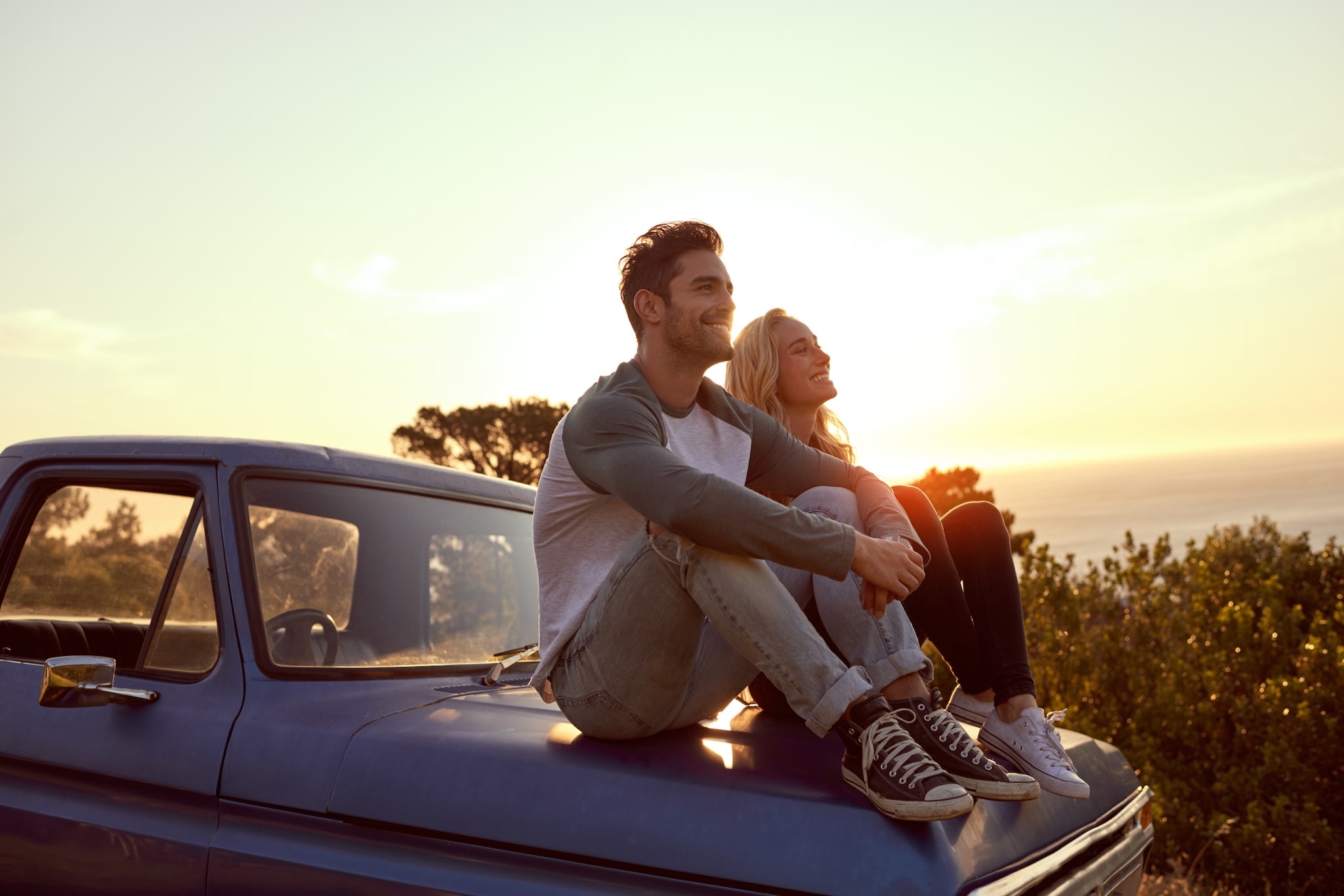 amex-insurances-reiseruecktrittsversicherung-fuer-individualreisen-paar-auto Ein junges Paar sitzt auf der Motorhaube eines Autos und genießt den Sonnenuntergang.