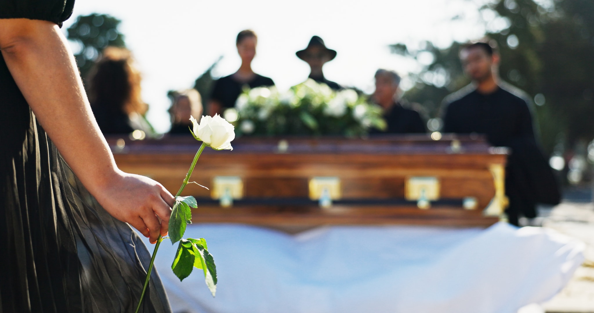 amex-insurances-reiseruecktrittsversicherung-tod-eines-angehoerigen-sarg-rose Eine in Schwarz gekleidete Person steht während einer Beerdigung mit einer weißen Rose vor einem Sarg, im Hintergrund weitere trauernde Personen.