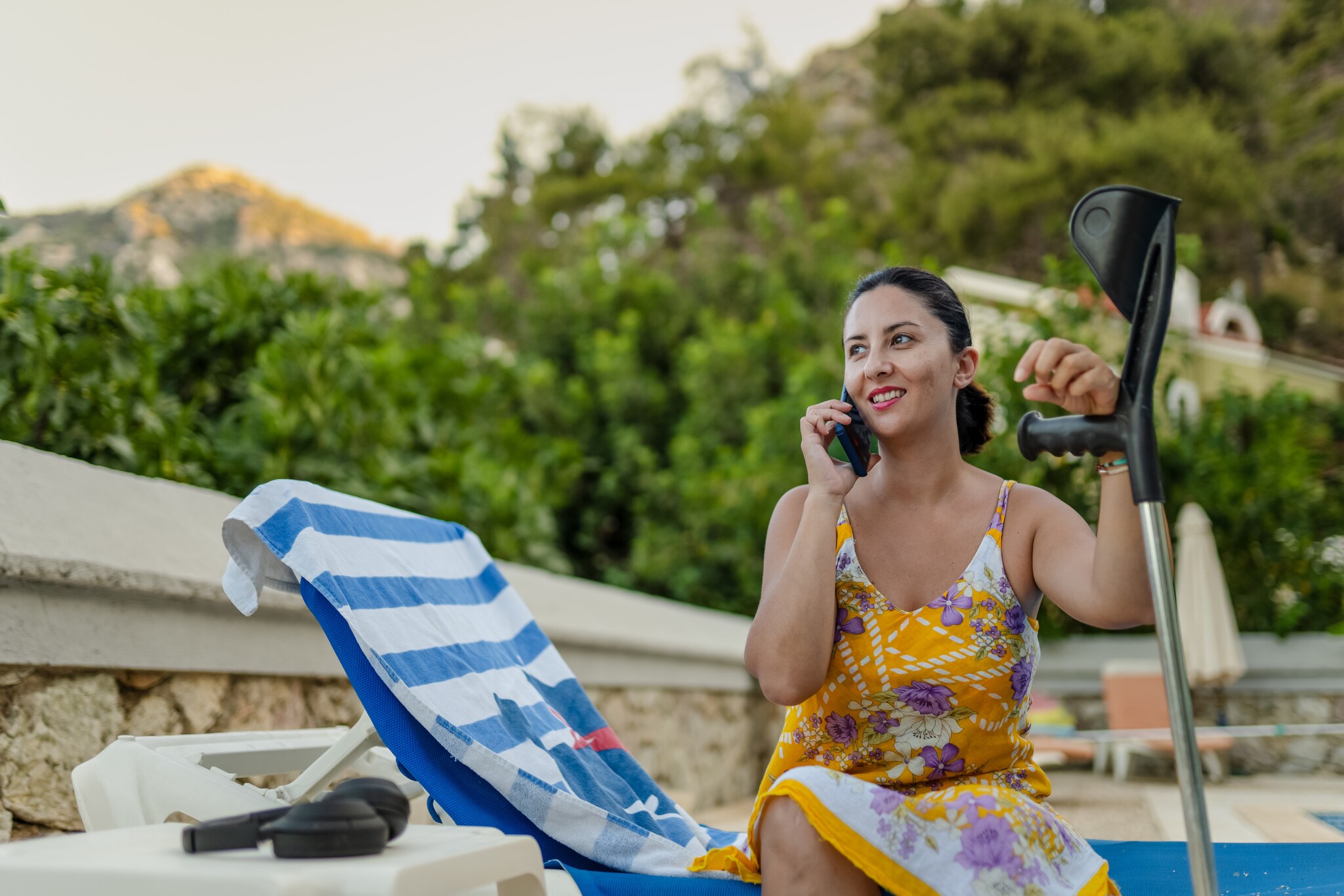 Eine Frau im Sommerkleid sitzt auf einer Sonnenliege am Pool und stützt ihren Arm auf einer Krücke ab.
