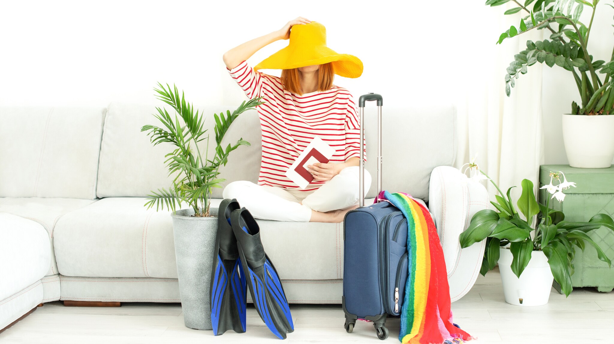 Eine Frau zieht sich ihren gelben Sonnenhut ins Gesicht. Sie hält ihren Reisepass in der Hand, neben ihr stehen Taucherflossen und Koffer.