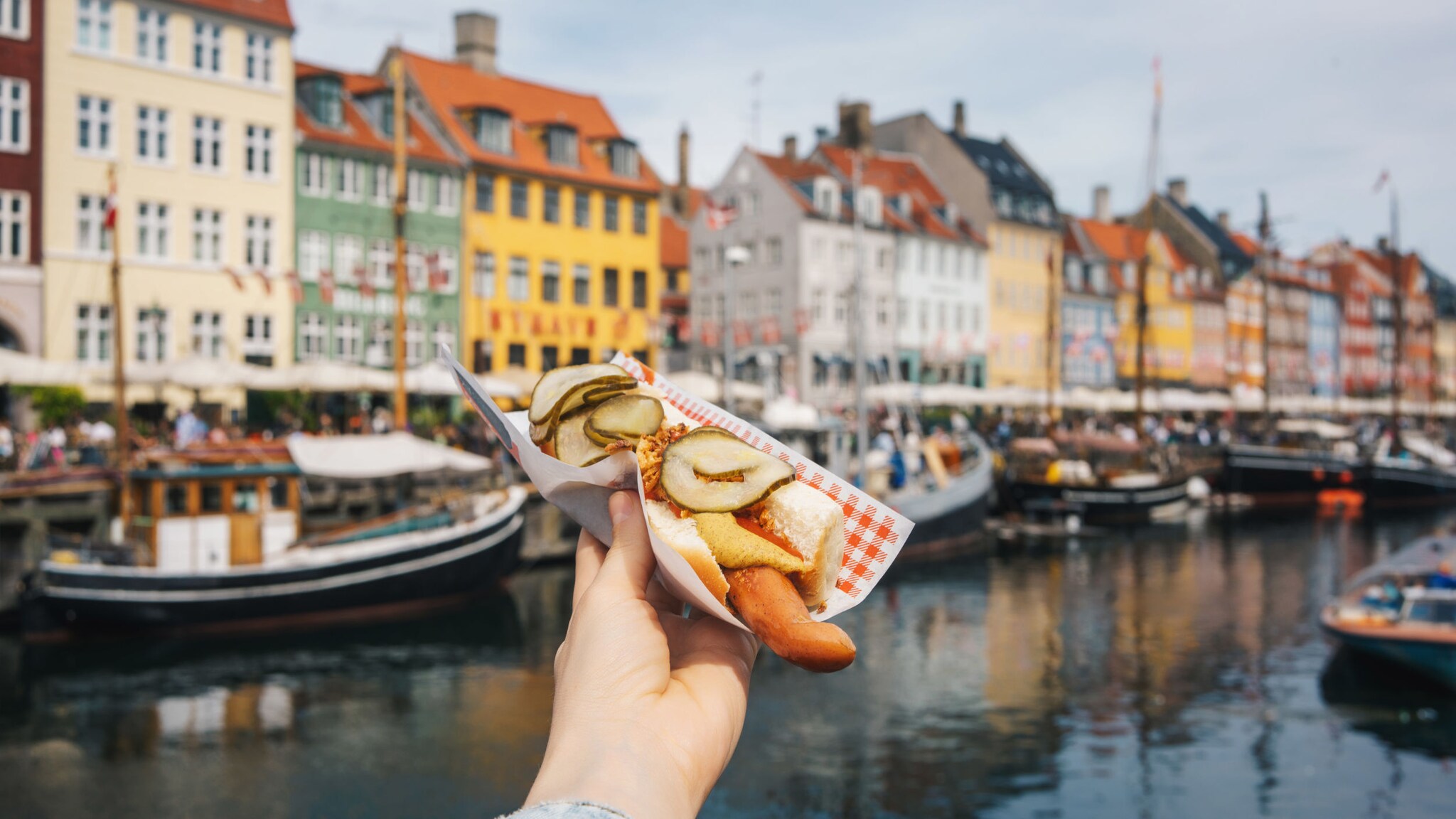 Ein Hotdog mit Gurken wird von einer Hand hochgehoben an einem Hafenbecken mit Holzbooten vor bunten Häusern.