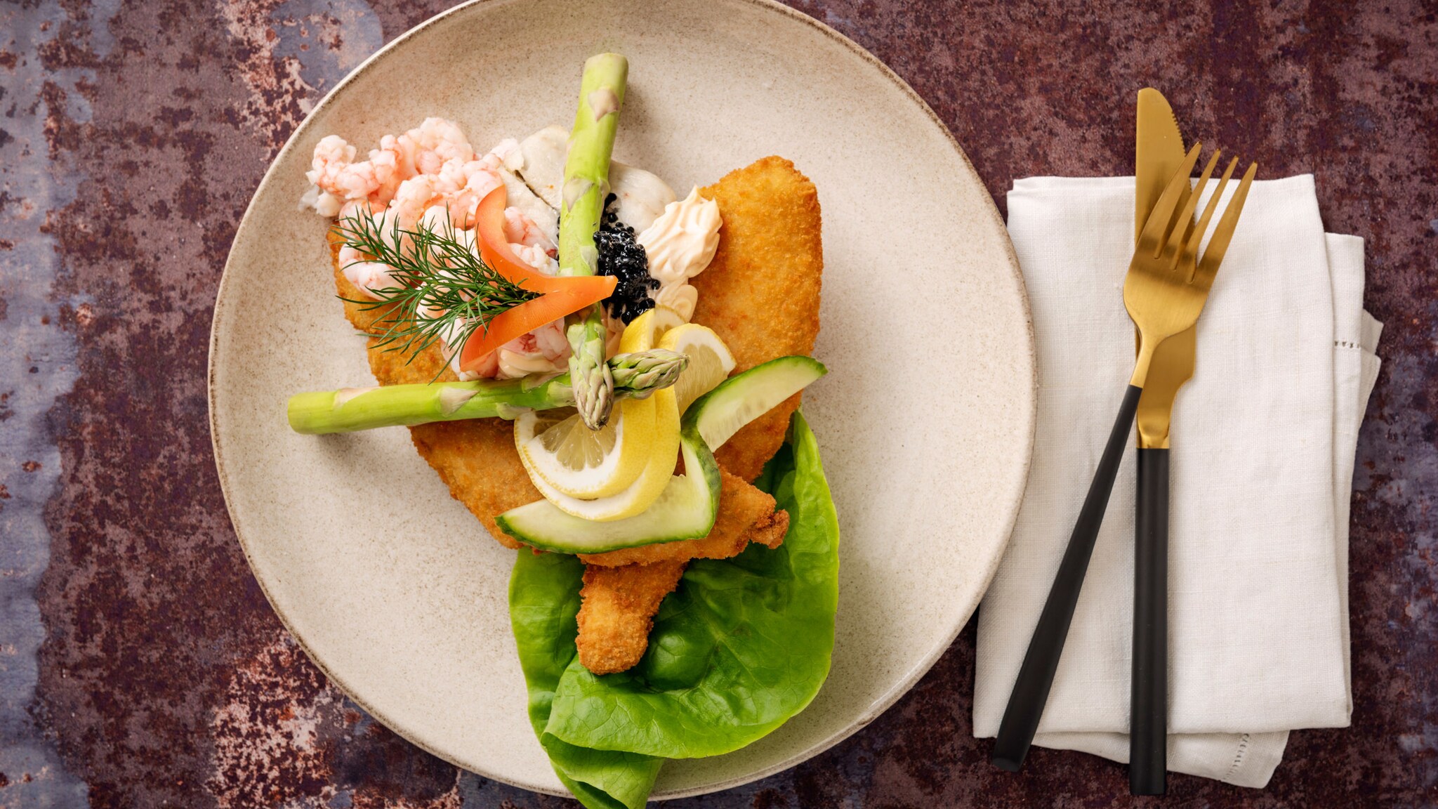 Aufsicht eines kunstvoll angerichteten Gerichtes mit zwei panierten Schollenfilets, Krabben und Salatbeilage auf einem weißen Teller, daneben elegantes Besteck auf einer weißen Serviette.