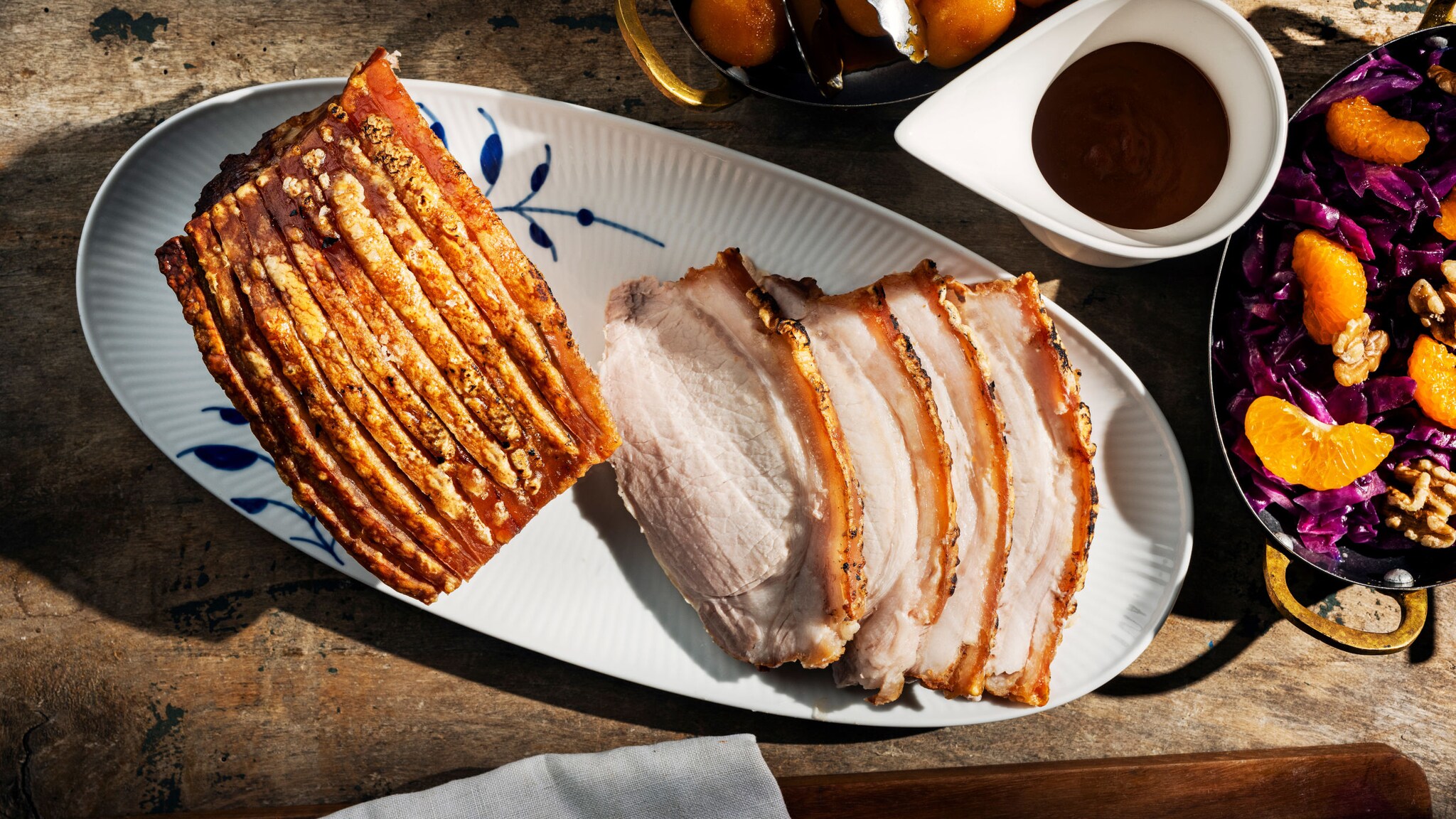 Aufsicht eines Schweinebratens mit tranchierten Scheiben auf einem Servierteller, daneben Schalen mit brauner Soße und Beilagen, auf einem Holztisch.