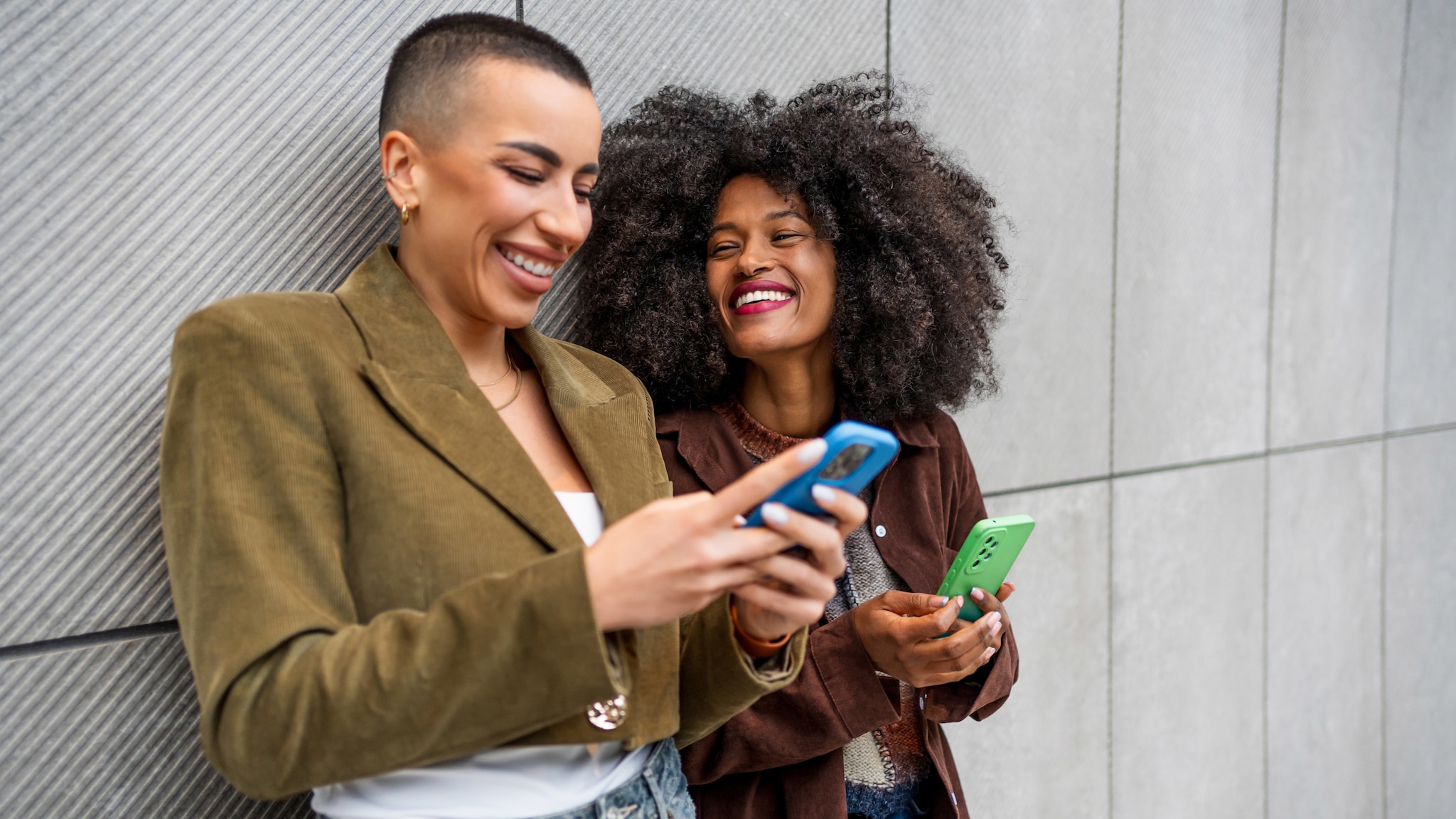 Zwei modern gekleidete Frauen mit Smartphones in den Händen lehnen lachend zusammen an einer Betonwand.