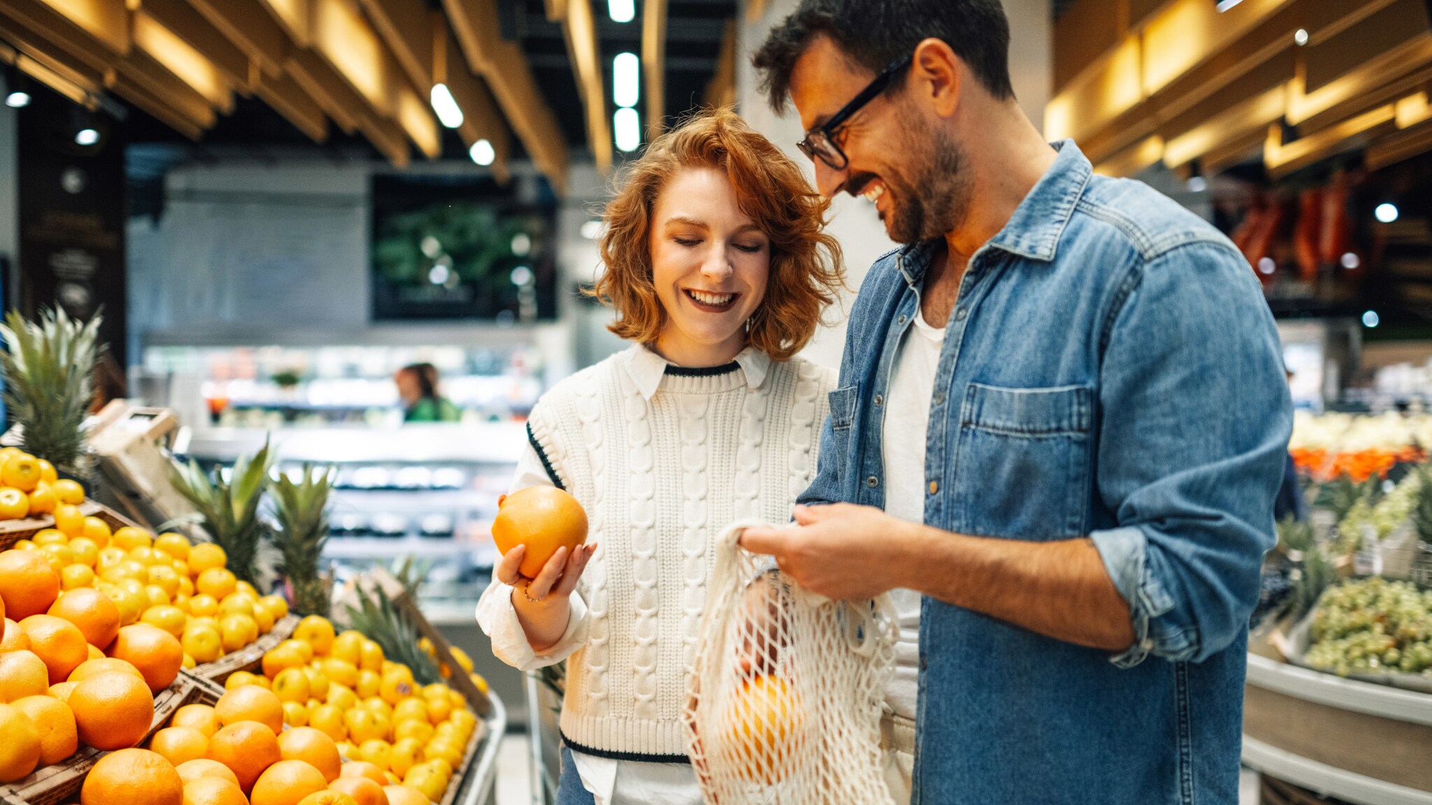 Eine Frau und ein Mann stehen lächelnd beieinander mit Orangen in Händen an einer Obsttheke in einem Supermarkt.