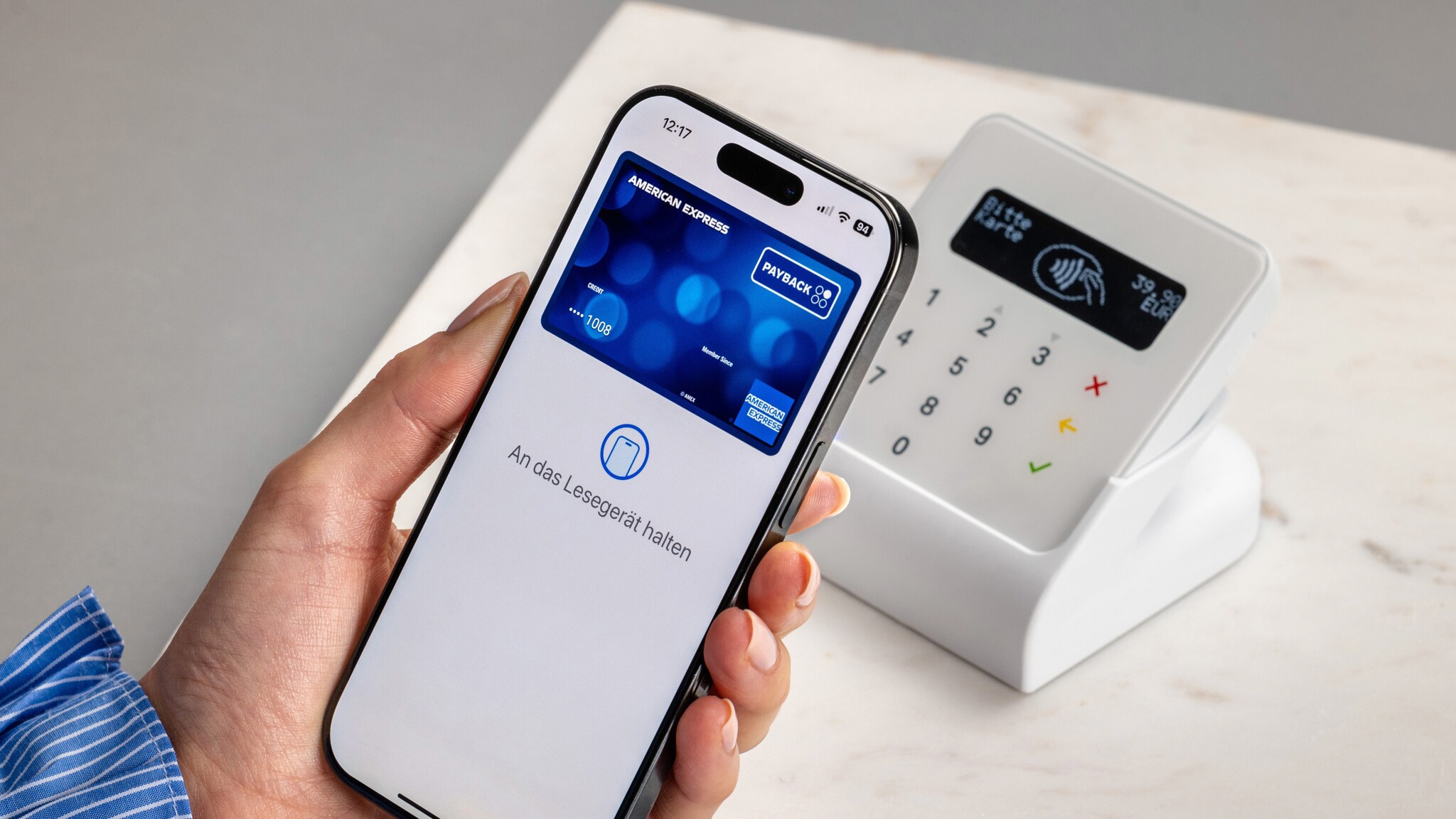 Kontaktlose Zahlung per Smartphone mit blauer PAYBACK American Express Kreditkarte auf dem Display in der Hand einer Person an einem Kartenlesegerät auf einem weißen Tisch.