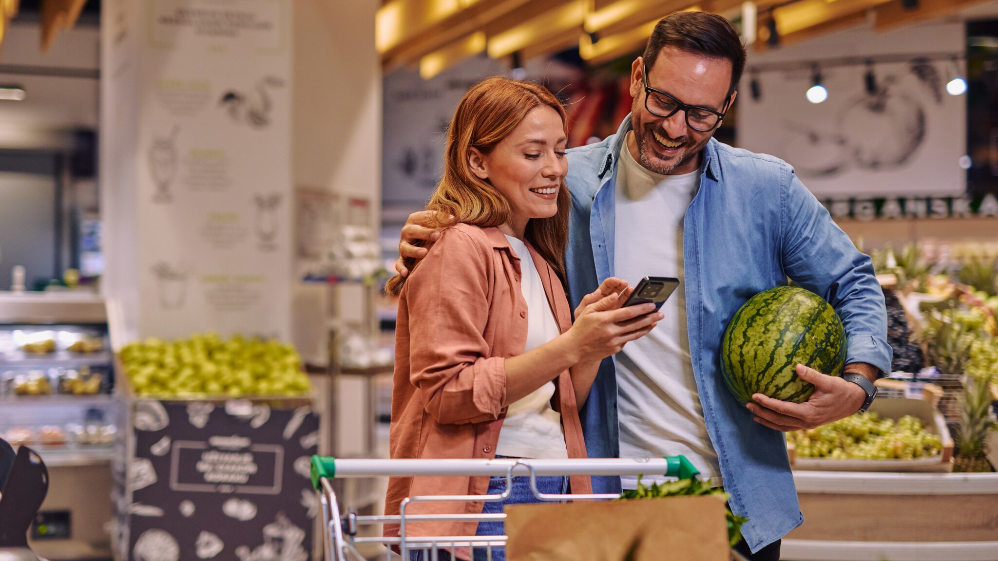 Eine Frau und ein Mann stehen eng beieinander in der Obstabteilung eines Supermarktes und schauen lächelnd auf ein Smartphone in ihrer Hand, er hält dabei eine Wassermelone im Arm.