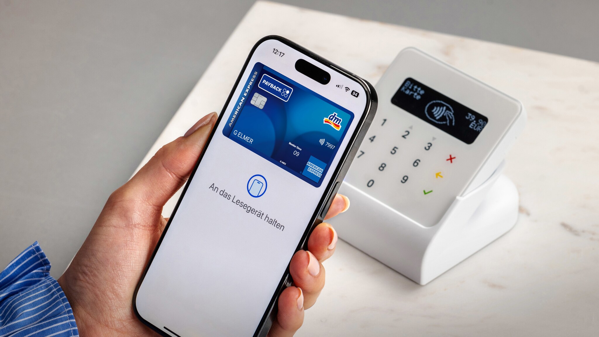 Kontaktlose Zahlung per Smartphone mit blauer dm PAYBACK American Express Kreditkarte auf dem Display in der Hand einer Person an einem Kartenlesegerät auf einem weißen Tisch.