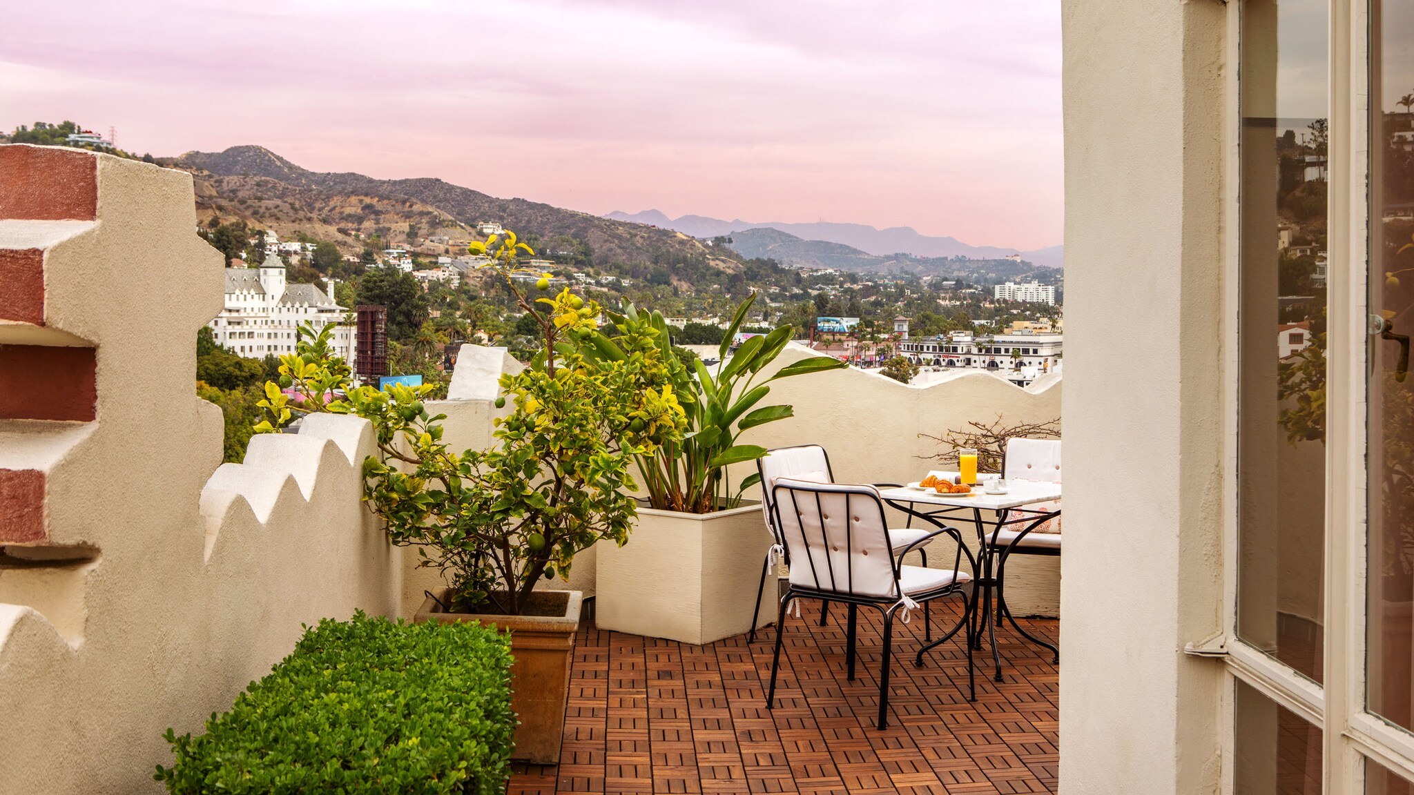 Umlaufende Terrasse mit eingedecktem Frühstückstisch mit Blick auf Hügellandschaft in Los Angeles unter rosa gefärbtem Himmel.