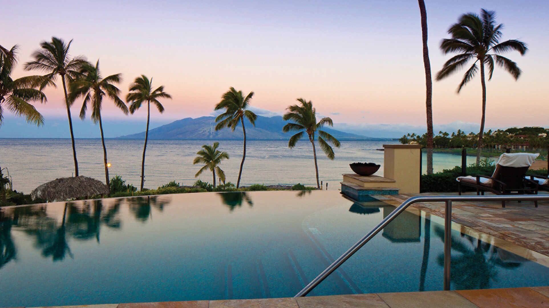 Infinity-Pool auf einer luxuriösen Hotelterrasse mit Palmen am Meer mit Blick auf eine Vulkaninsel im Abendlicht.