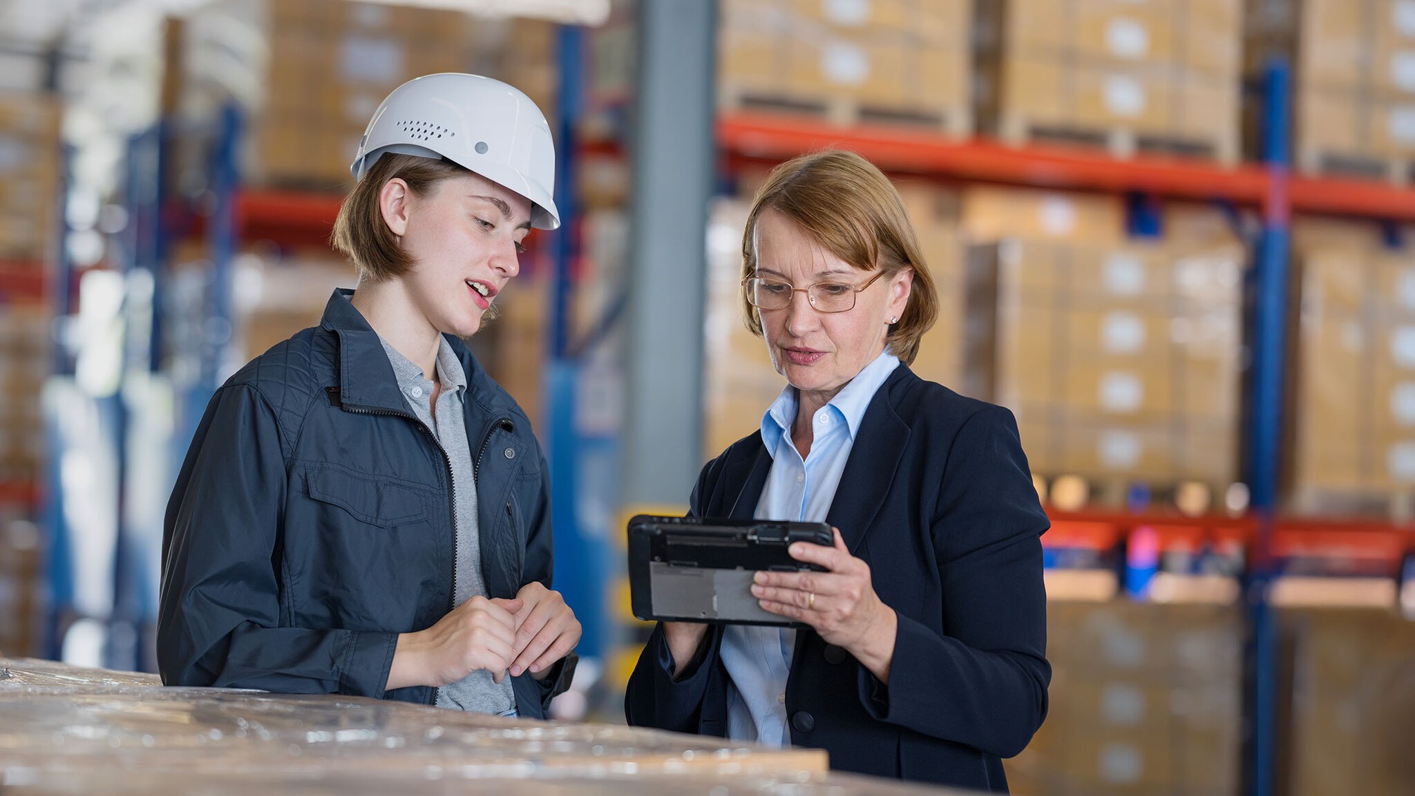 Eine Frau mit Helm und eine Frau in Businesskleidung stehen in einem Lager und schauen sich etwas auf einem Tablet an