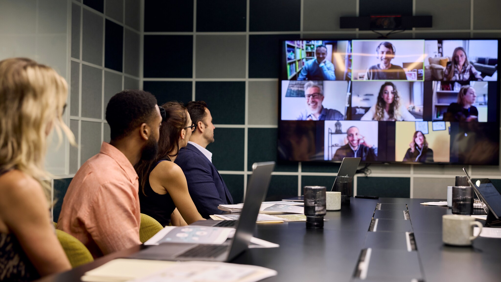 Mehrere Personen sitzen an einem Konferenztisch in einem Büro und blicken auf einen Monitor mit Personen auf unterschiedlichen Kacheln während eines Meetings.