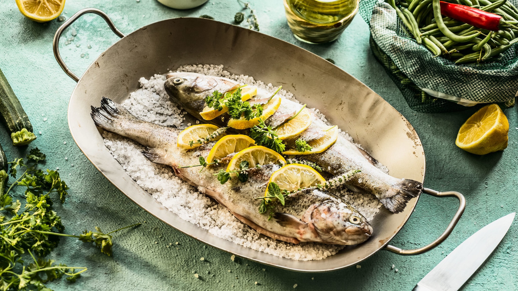Zwei mit Salz und Zitronenscheiben bedeckte Forellen in einer Backform auf einem Tisch mit Kochzutaten.