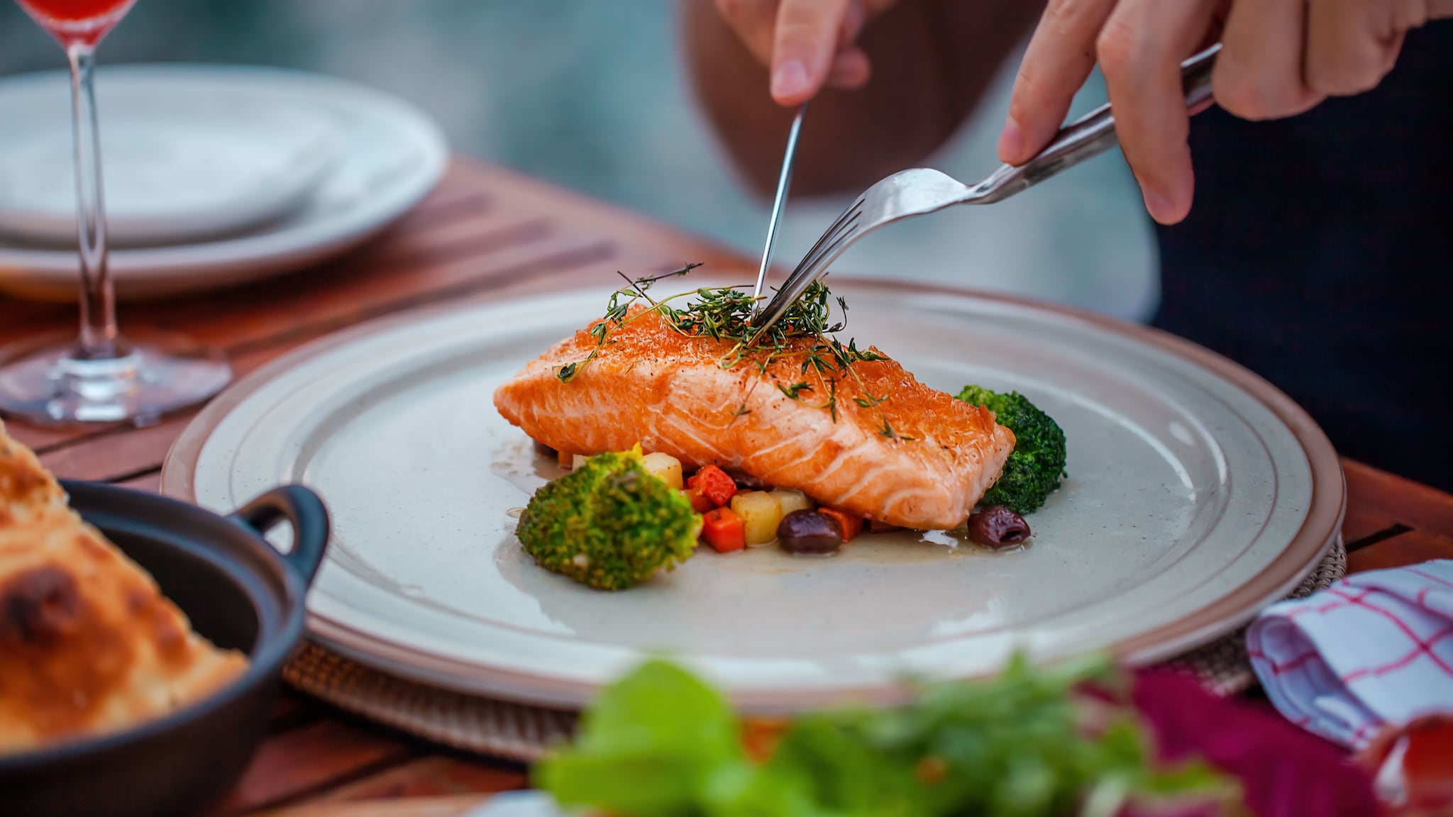 amex-magazin-fisch-essen-lachsfilet Messer und Gabel in den Händen einer Person an einem gebratenen Lachsfilet, angerichtet auf Gemüse, auf einem weißen Teller auf einem Tisch.
