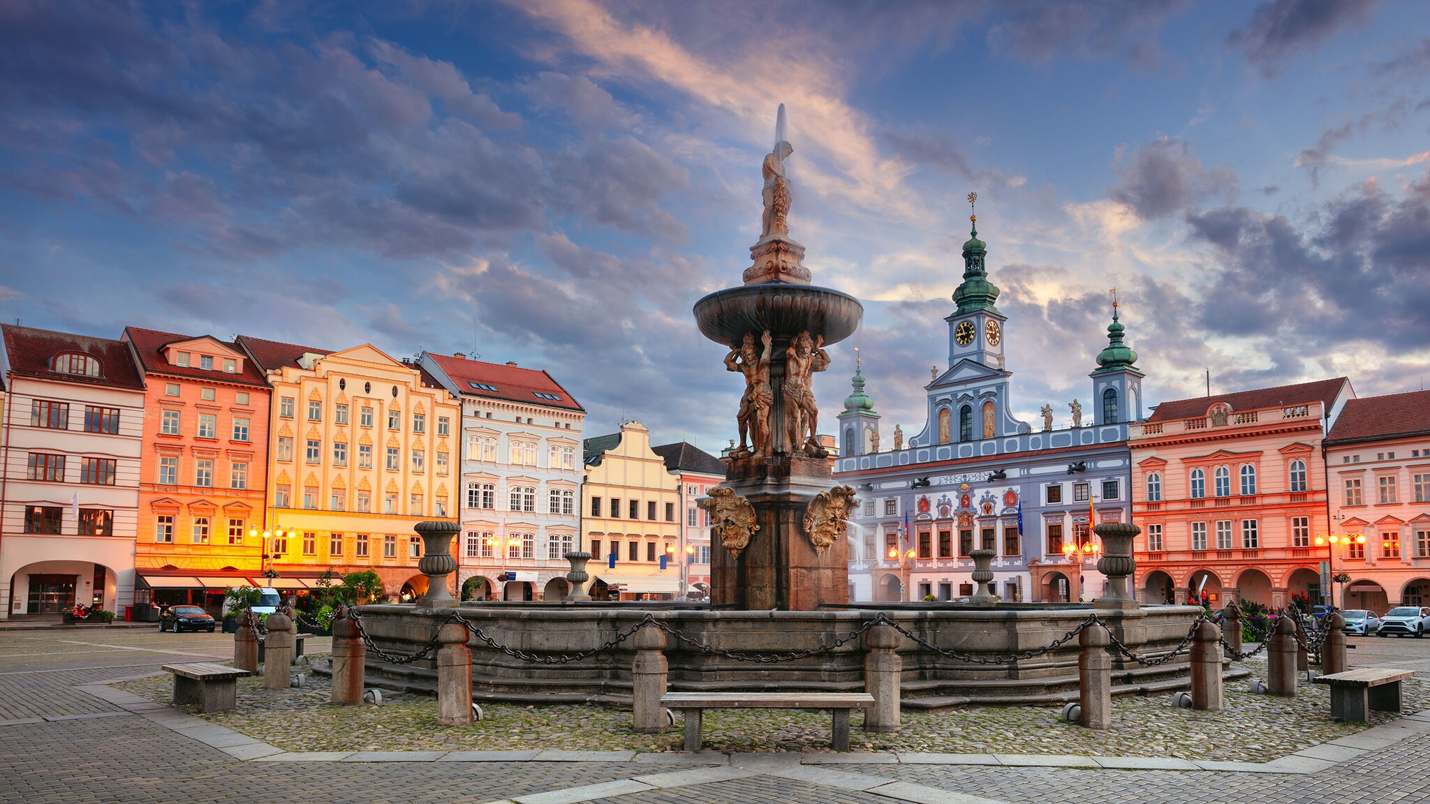 Auf einem Platz steht ein verzierter Brunnen vor bunten, historischen Häusern. 