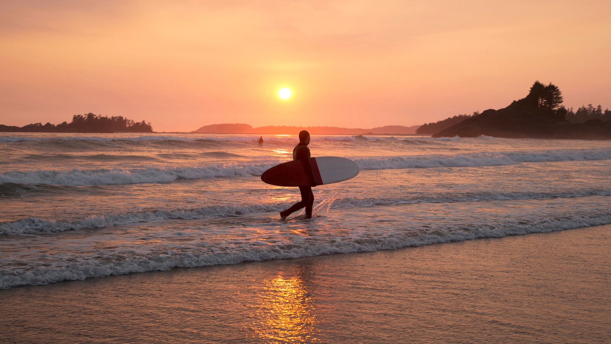 Ein Mann geht mit Surfbrett unter dem Arm im welligen Wasser an einem Sandstrand entlang bei Sonnenuntergang mit rosa gefärbtem Himmel.