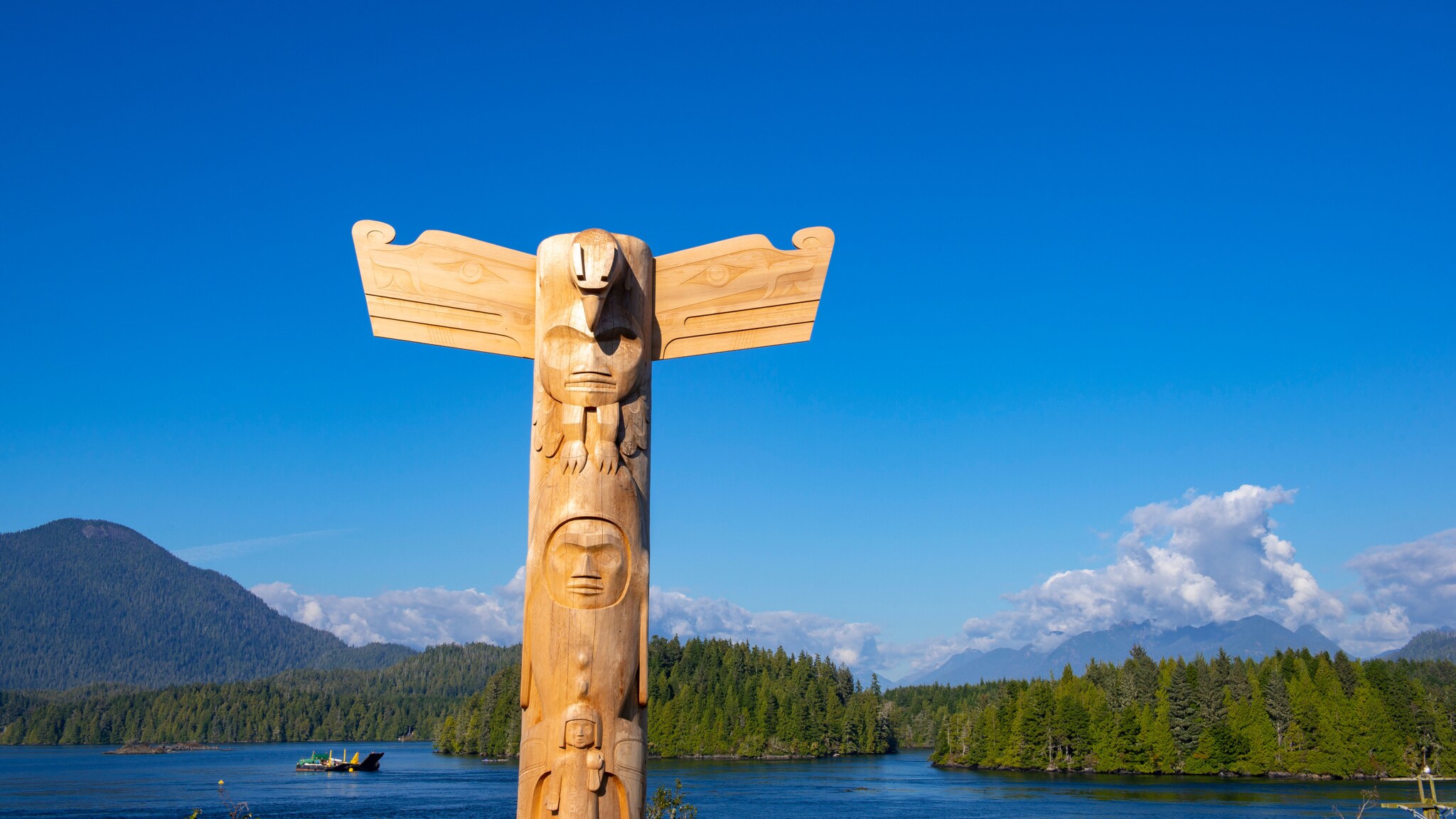 Ein Totempfahl vor einem Gewässer mit Waldgebiet unter blauem Himmel.