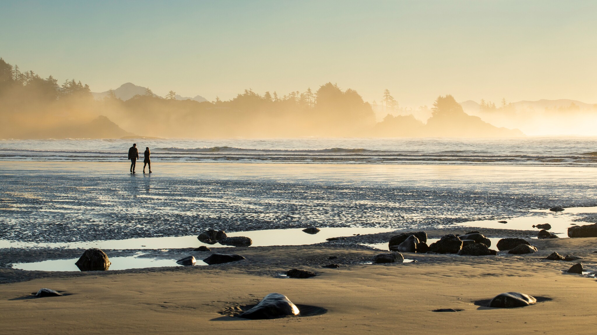 Zwei Personen laufen im flachen Wasser an einem weiten Sandstrand im Nebel.