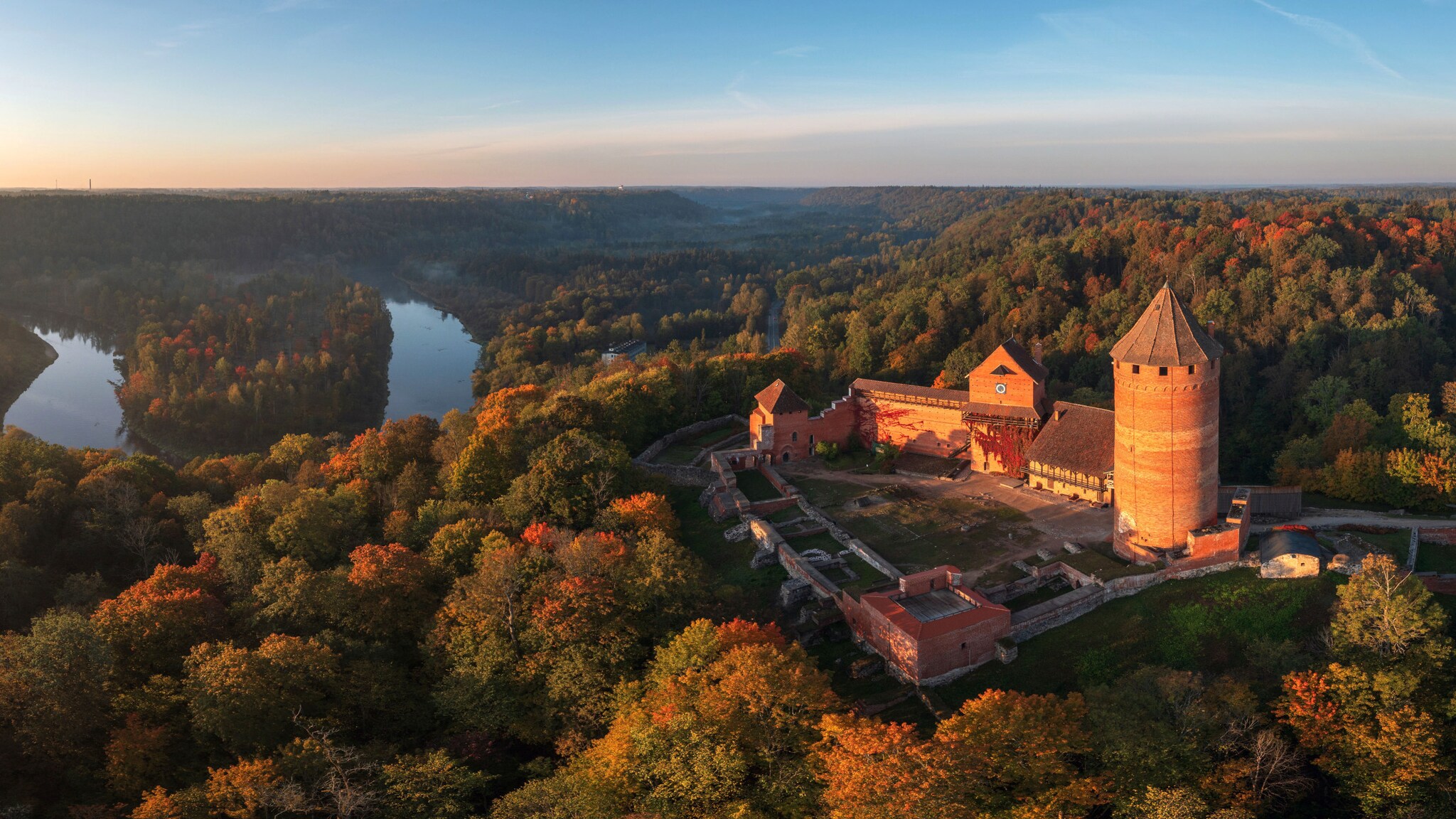 Rote Burganlage in einem hügeligen Waldgebiet mit Flusstal im warmen Sonnenlicht.
