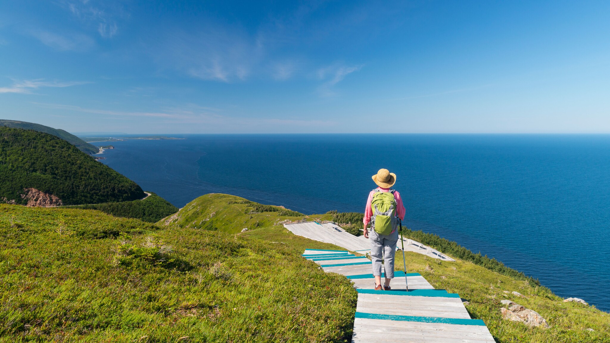 Rückansicht einer Person mit Sonnenhut, Rucksack und Wanderstock auf einem Holzpfad auf einem grünen Berg an einer Küste unter blauem Himmel.