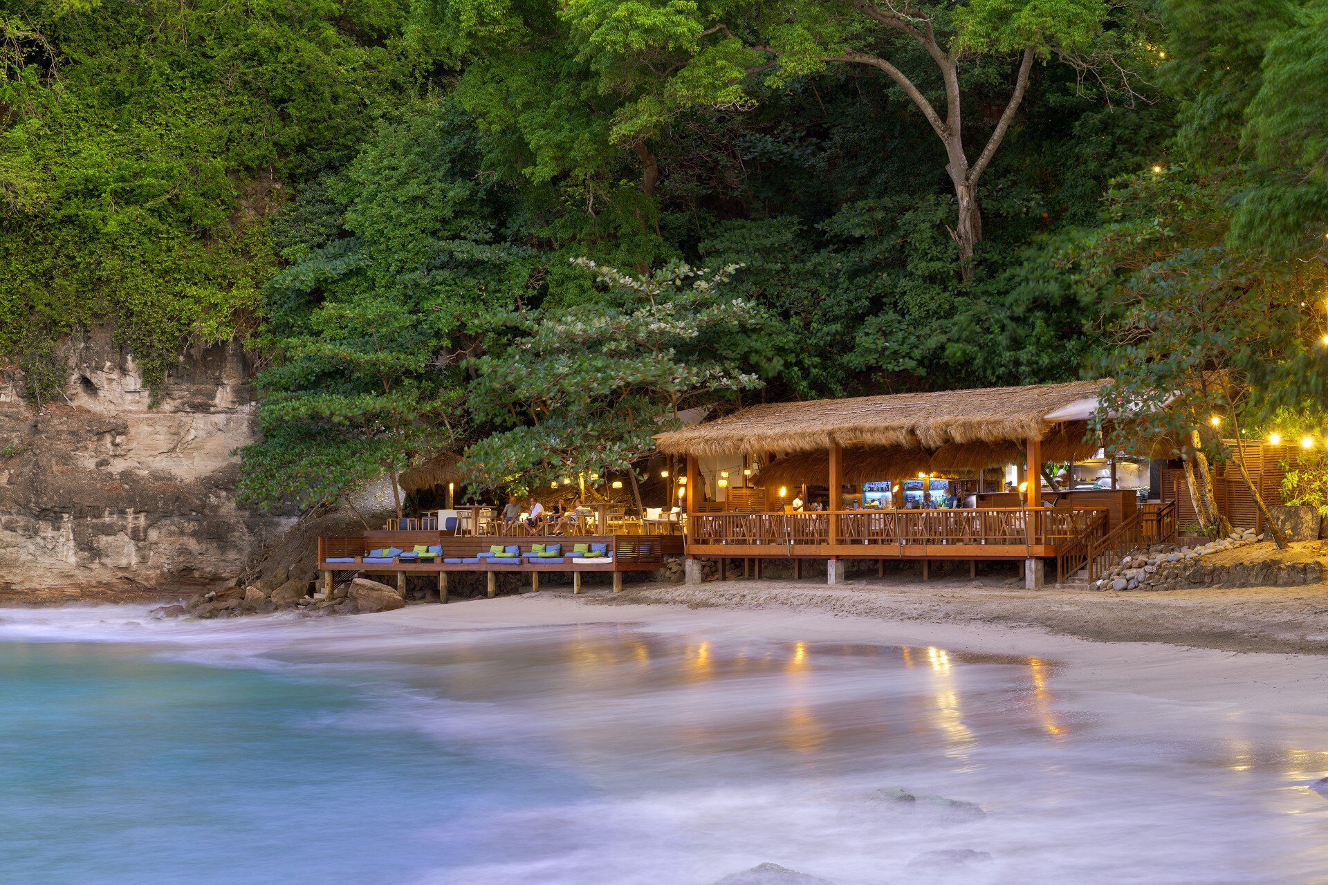 Strandrestaurant mit Strohdach auf einer Holzterrasse direkt am Meer, umgeben von tropischer Vegetation und Felsen.