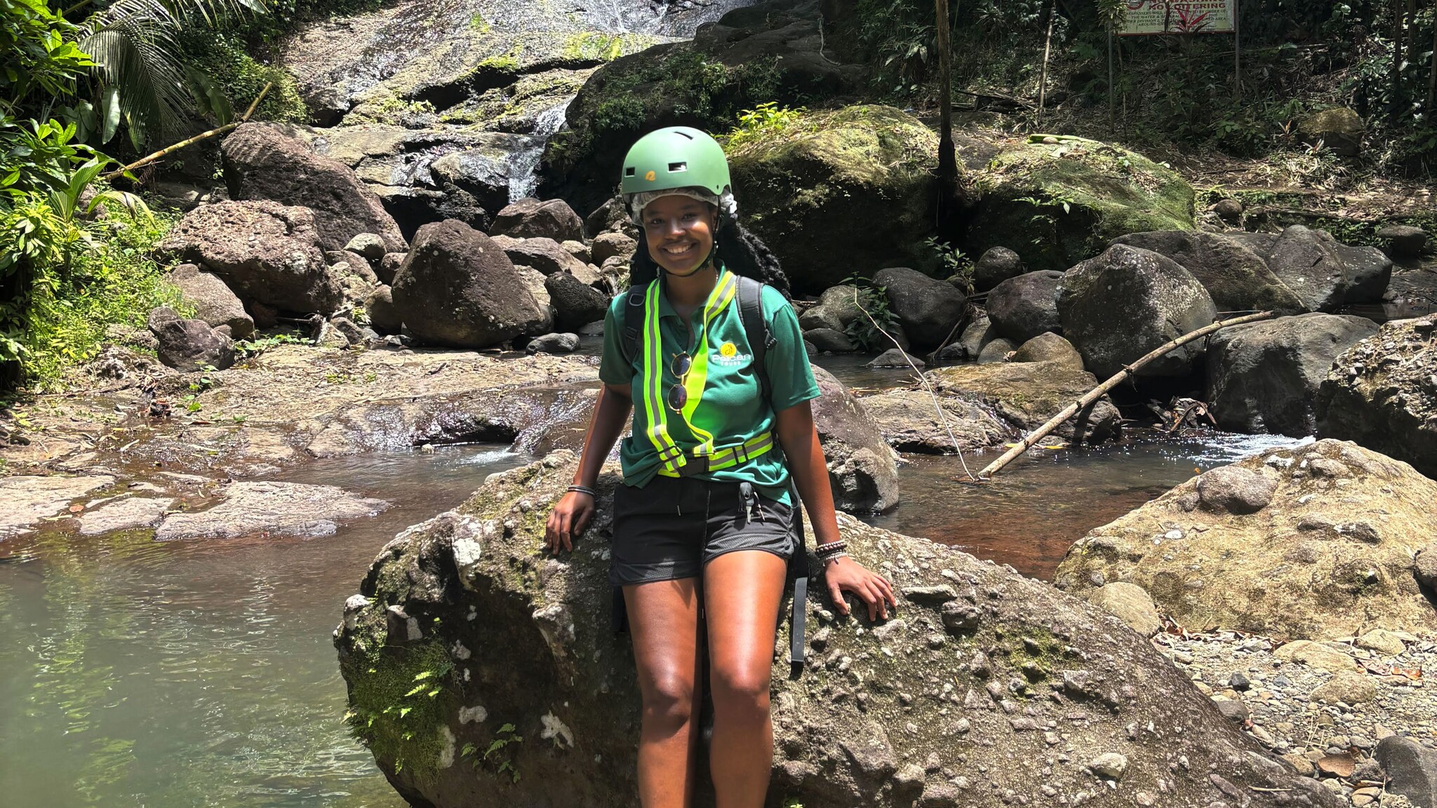 Person mit Helm sitzt auf einem Felsen vor einem kleinen Wasserfall in dichtem, tropischem Wald.